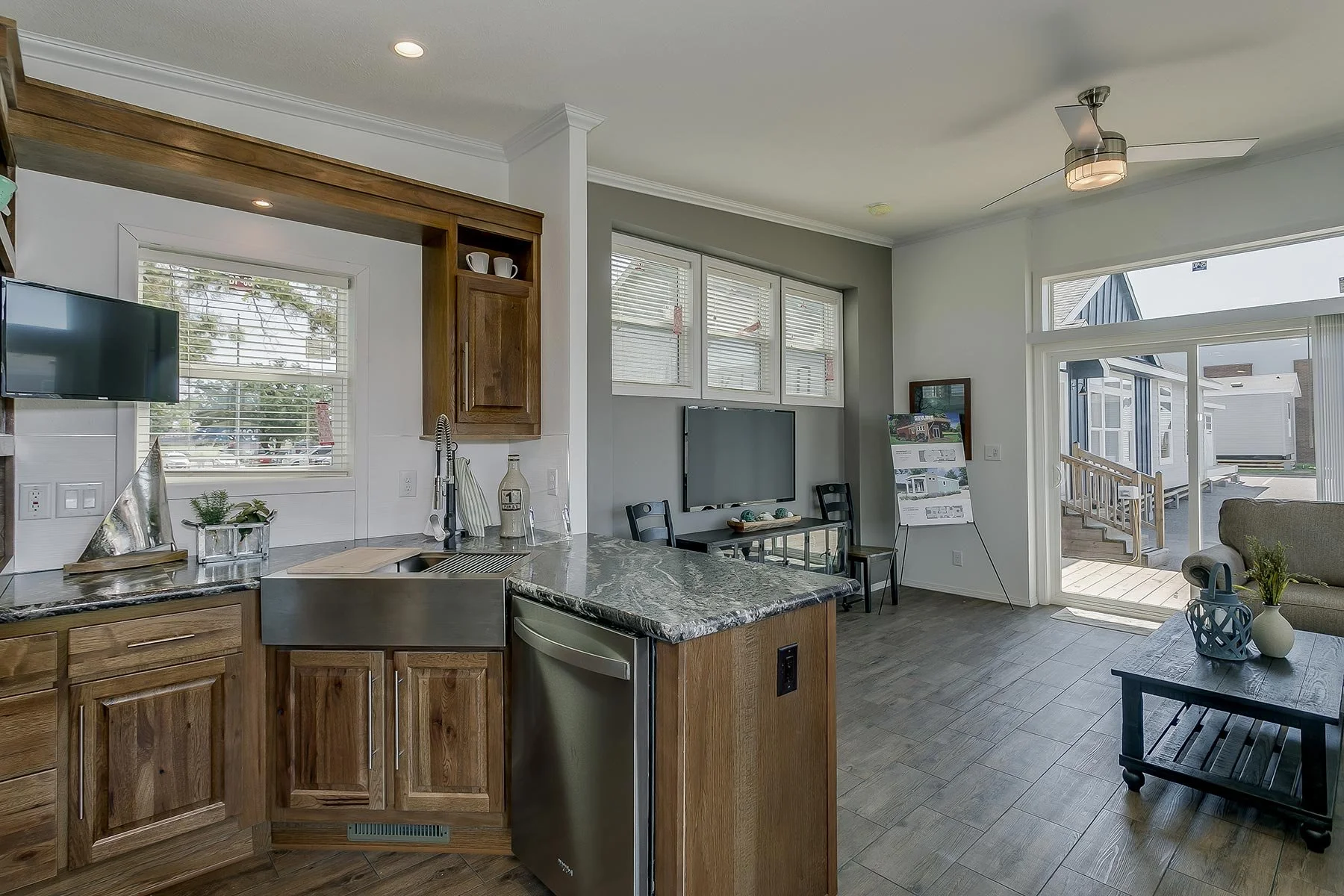 Open-concept kitchen and living room with wood cabinets, granite countertops, stainless steel sink, small TV, windows with blinds, and sliding glass door leading outside to a porch with steps and neighboring houses.