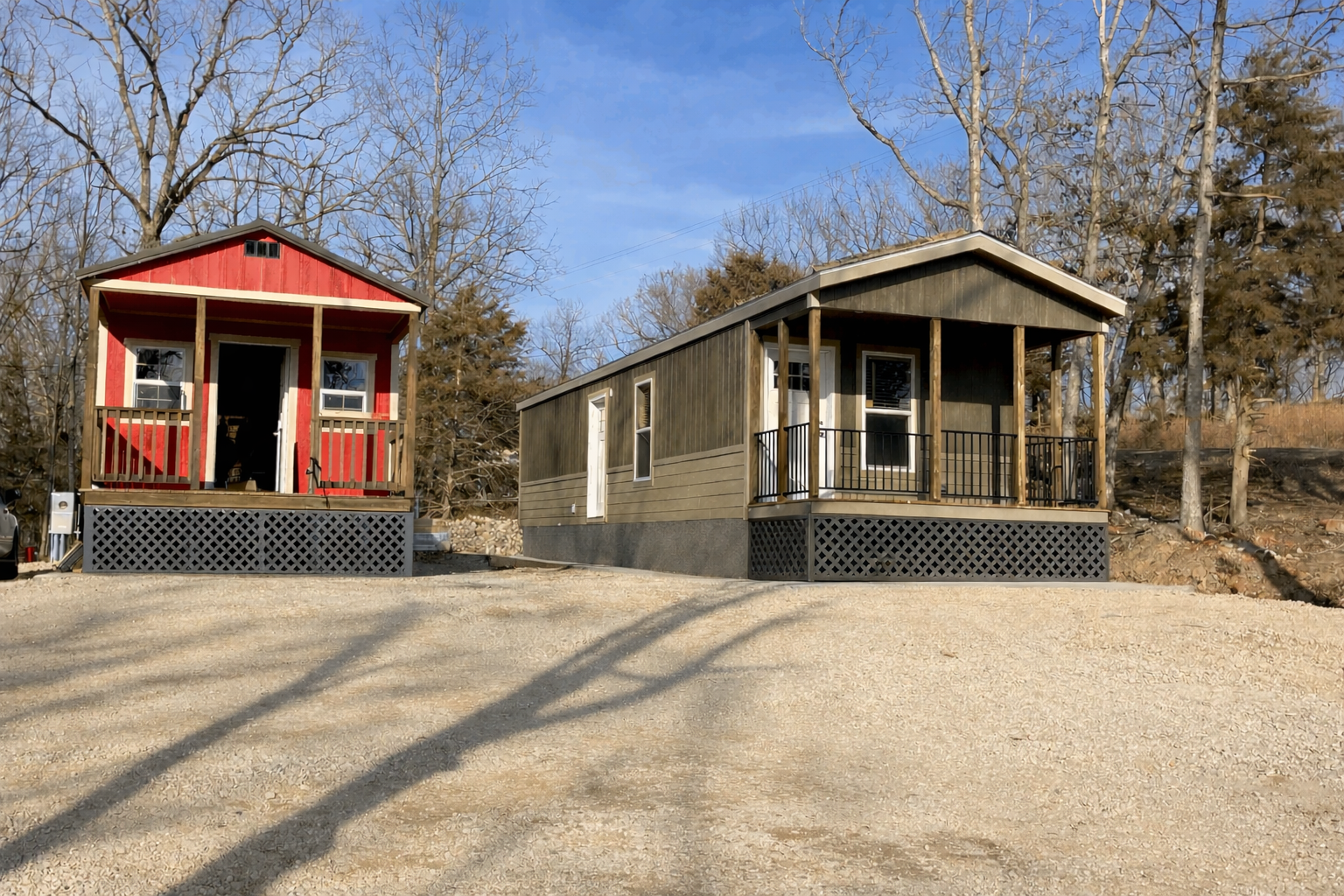 Two tiny houses on gravel ground, one painted red and the other painted dark green, with trees and a blue sky in the background.