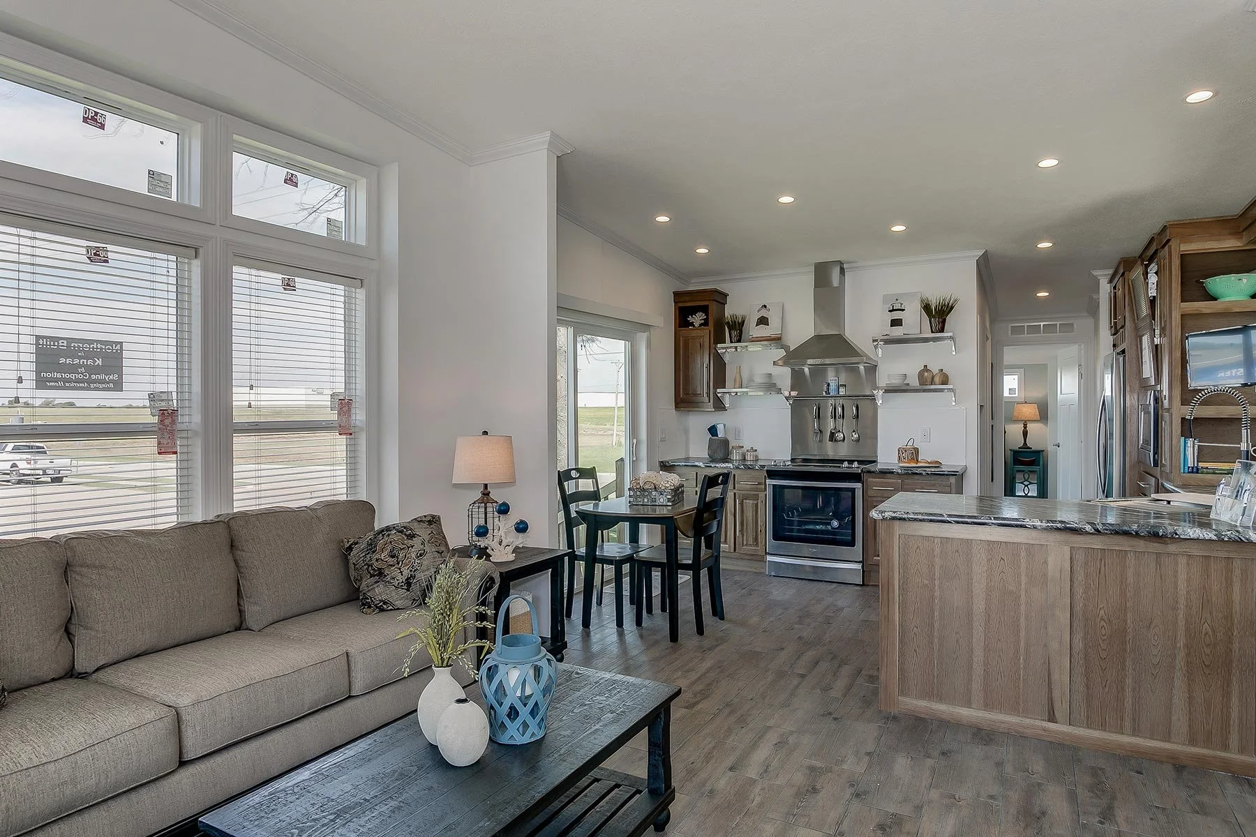 Open-concept living room and kitchen with large windows, beige sofa, coffee table with decorative vases, and kitchen with stainless steel appliances, wooden cabinets, and granite countertops.