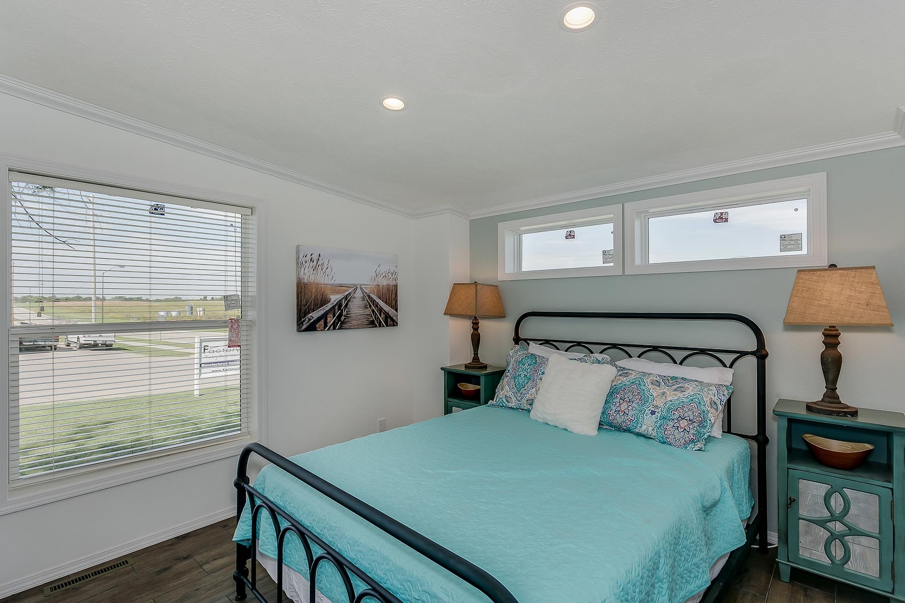 A bedroom with a black metal bed frame, turquoise bedding, and patterned pillows. There are two nightstands with lamps and a door with blinds opening to a grassy outdoor scene.