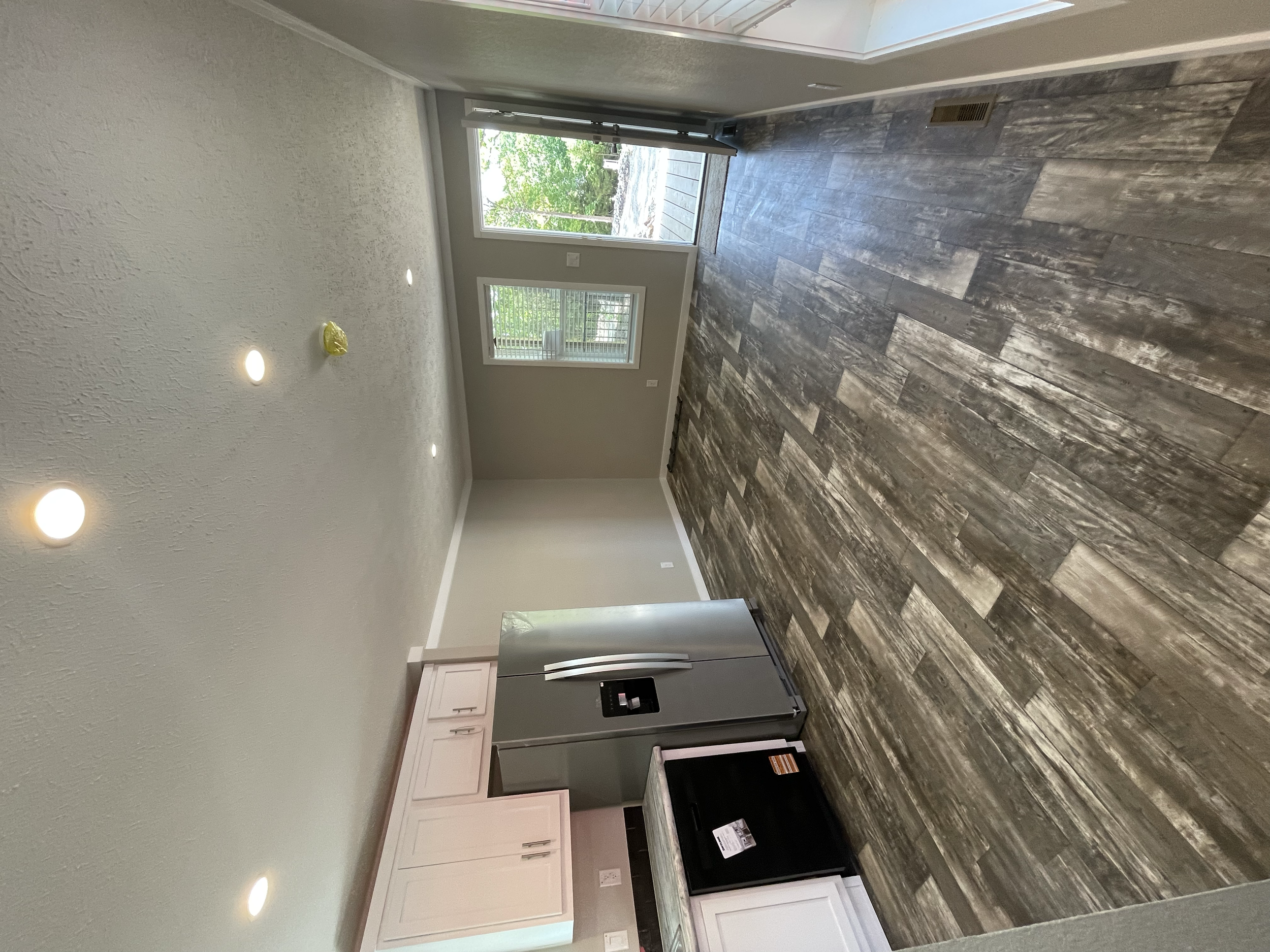 Inside view of a modern kitchen with white cabinets, a large stainless steel refrigerator, and dark wood flooring. There is a sliding glass door leading to an outdoor area with greenery.