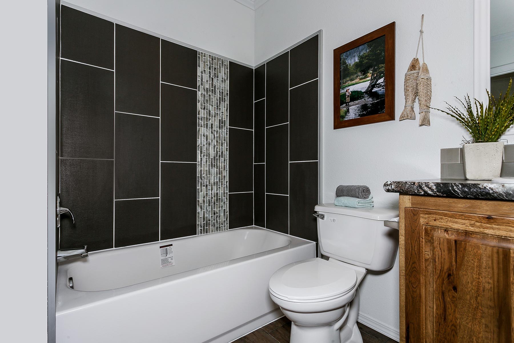 Bathroom with a white bathtub, black tiled shower wall with a vertical strip of small gray mosaic tiles, a white toilet with folded towels on top, a wooden vanity with a granite countertop, a potted plant, picture on the wall, and decorative fish wal