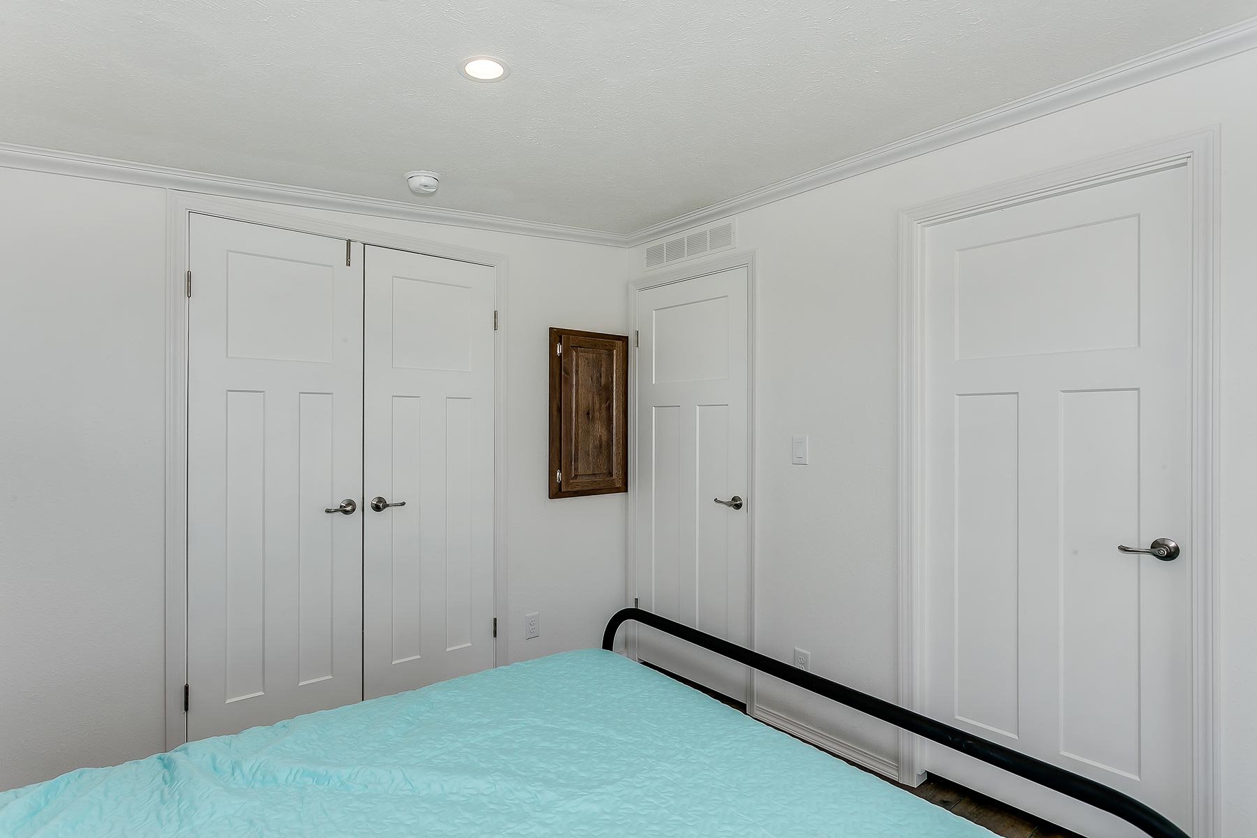 A bedroom with white walls, white doors, a bed with a light blue quilt, and a black bed frame. There is a small wooden cabinet mounted on the wall and two ceiling lights.