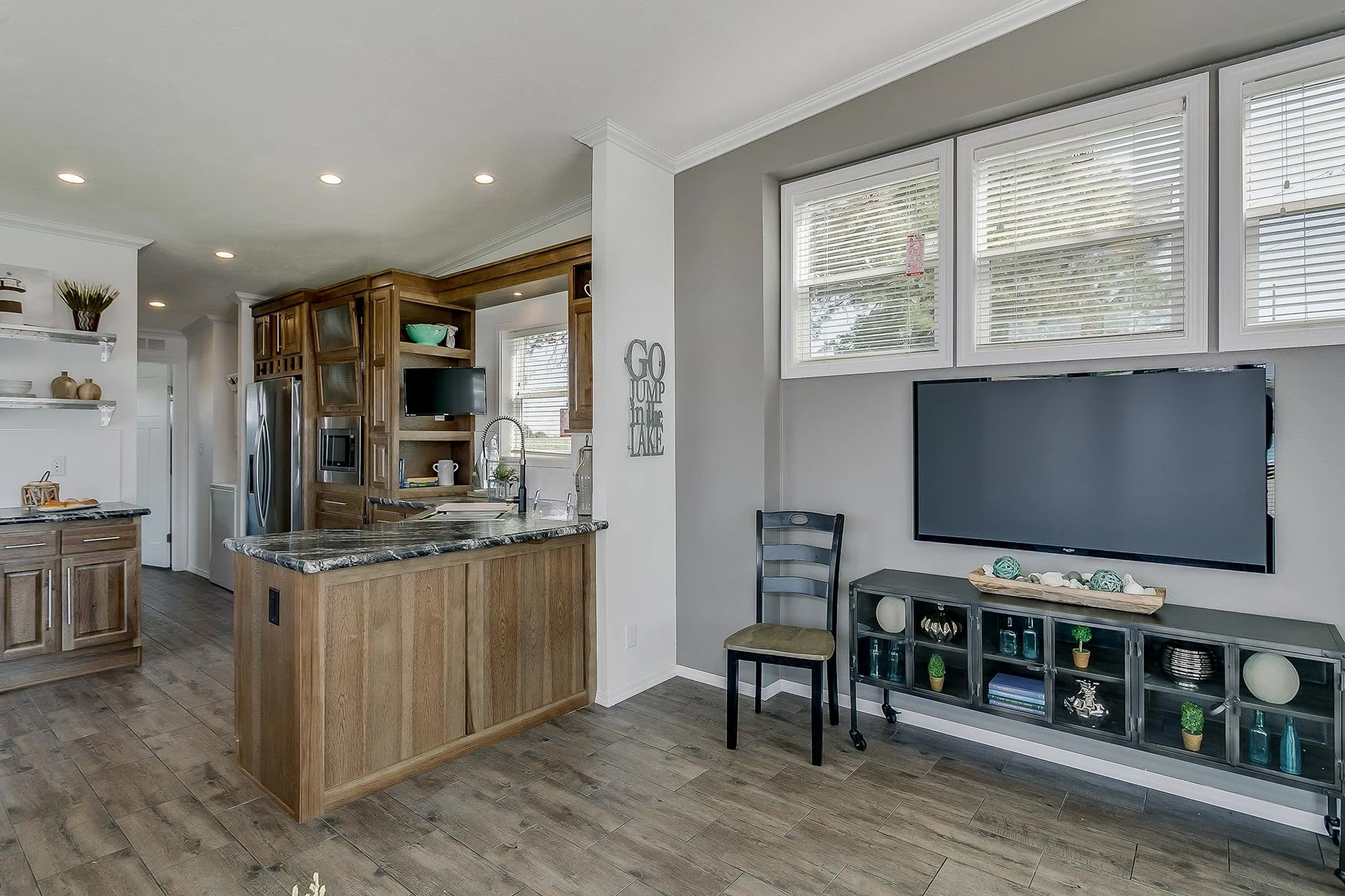 Interior view of a modern kitchen and living room area with wooden cabinets, a granite countertop, a wall-mounted flat-screen TV, and decorative items on open shelves.