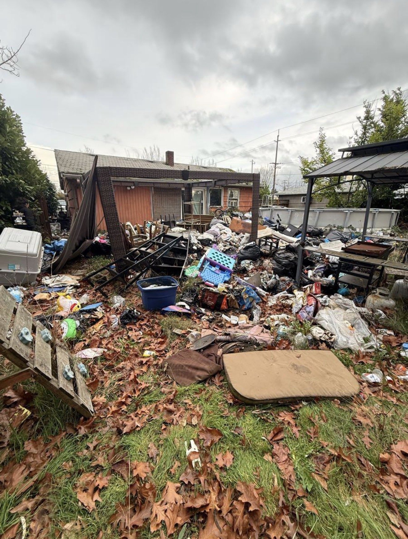 An outdoor scene with a house in the background, and the yard is filled with trash, including scattered debris, empty containers, broken furniture, and miscellaneous rubbish on a bed of fallen leaves.