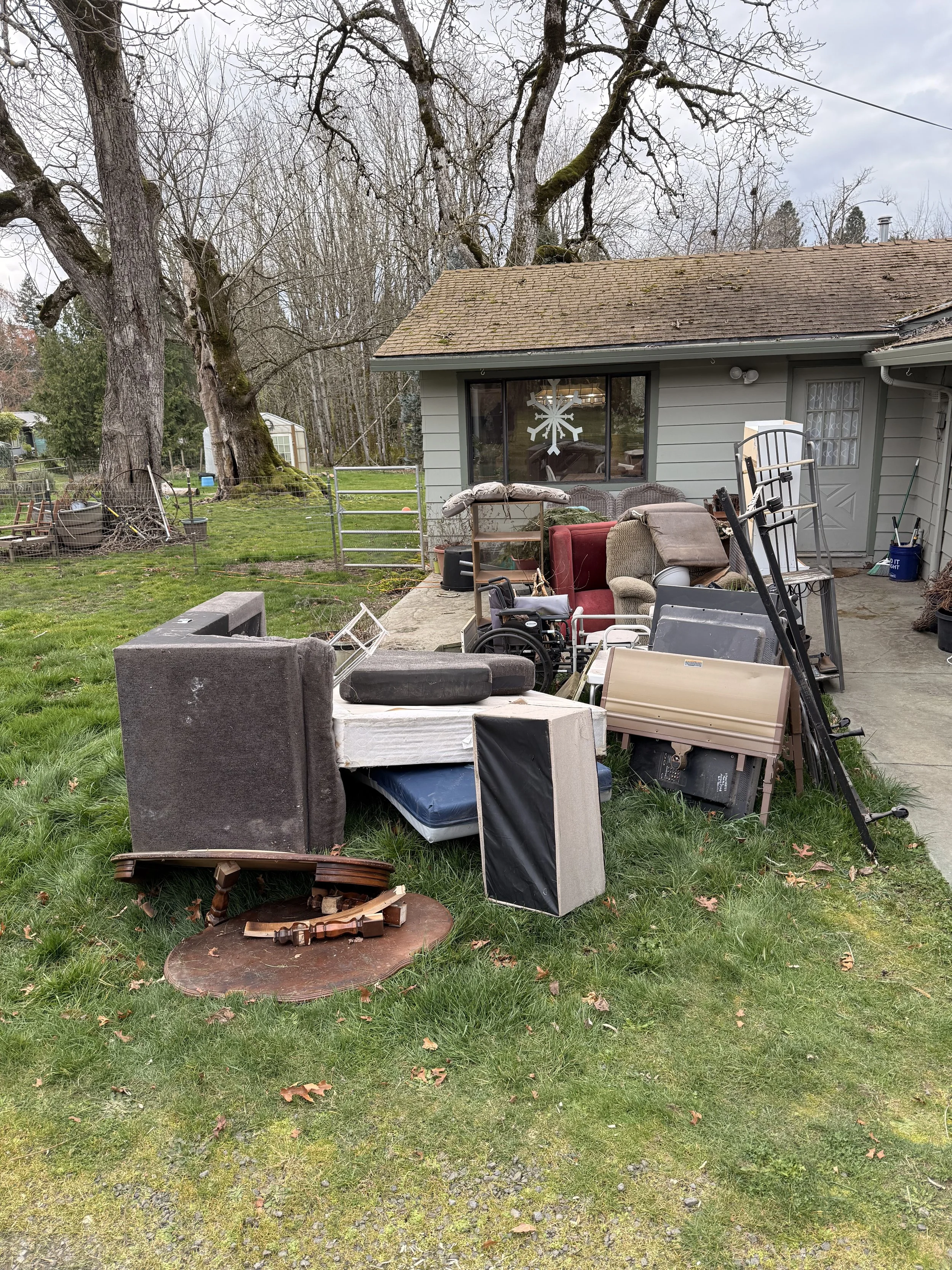 A backyard with various discarded furniture and household items scattered on the grass and patio, including a sofa, a wheelchair, a small table, a mirror, and miscellaneous debris, with a house in the background and leafless trees.