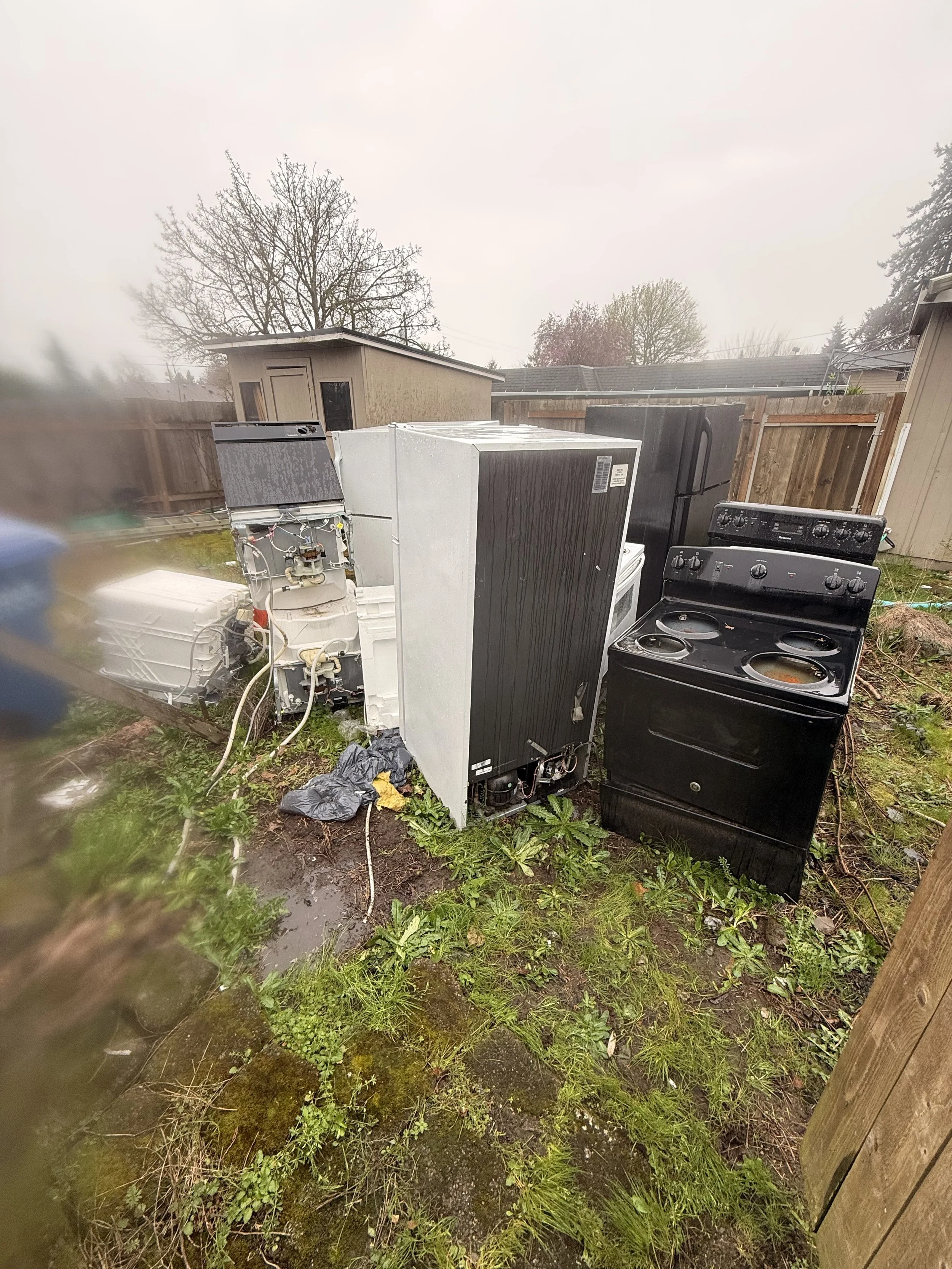 Disassembled and disorganized appliances, including a washing machine, a dryer, and a stove, placed outdoors in a backyard with grass, muddy patches, and weeds, surrounded by a wooden fence and trees.
