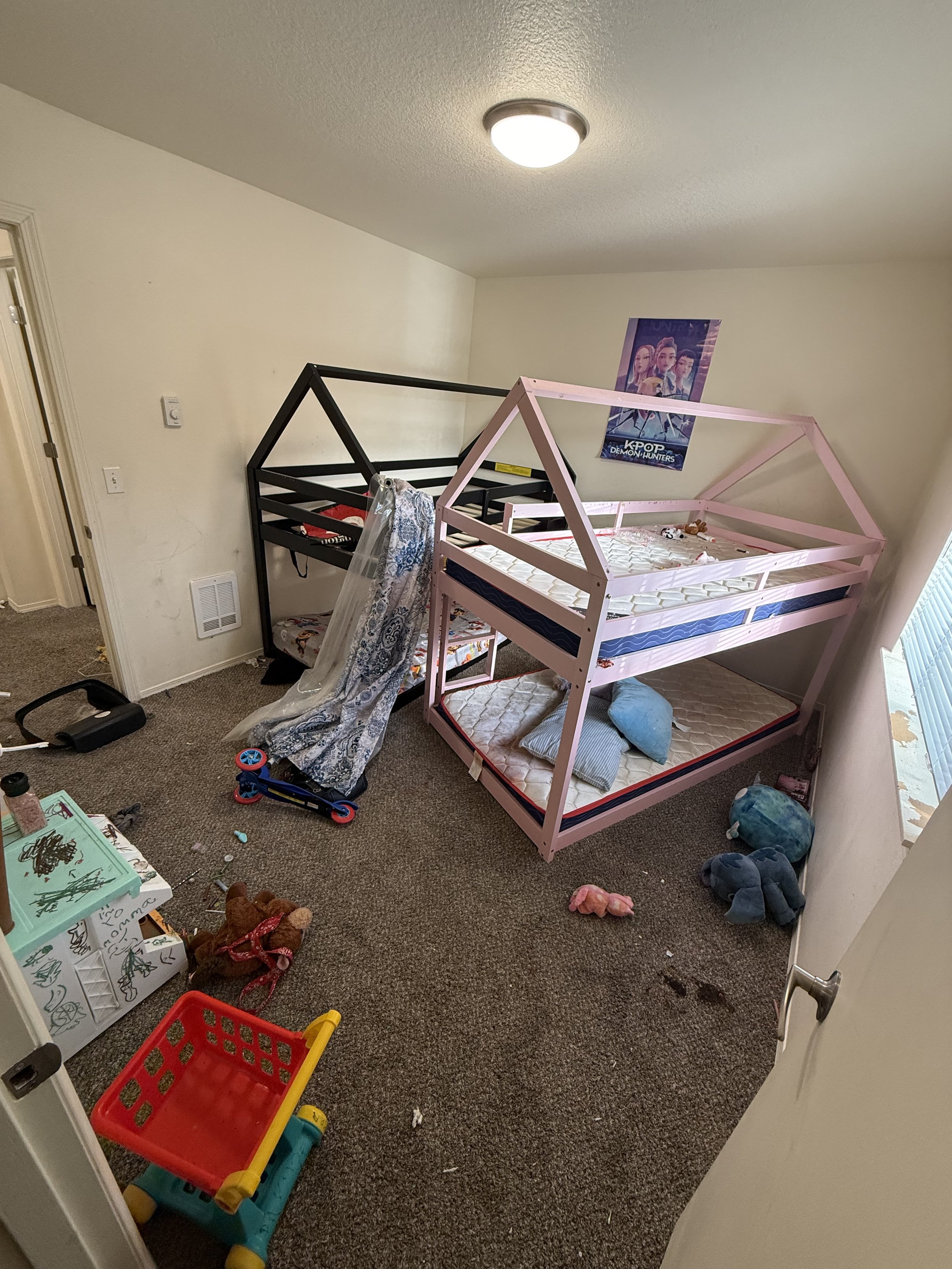 A messy children's bedroom with two bunk beds, one painted pink and the other black, both unmade. The room has toys and scattered items on the carpeted floor, including a red and yellow toy shopping cart, stuffed animals, and art supplies. There is a poster of K-pop artists on the wall and a window with closed blinds.