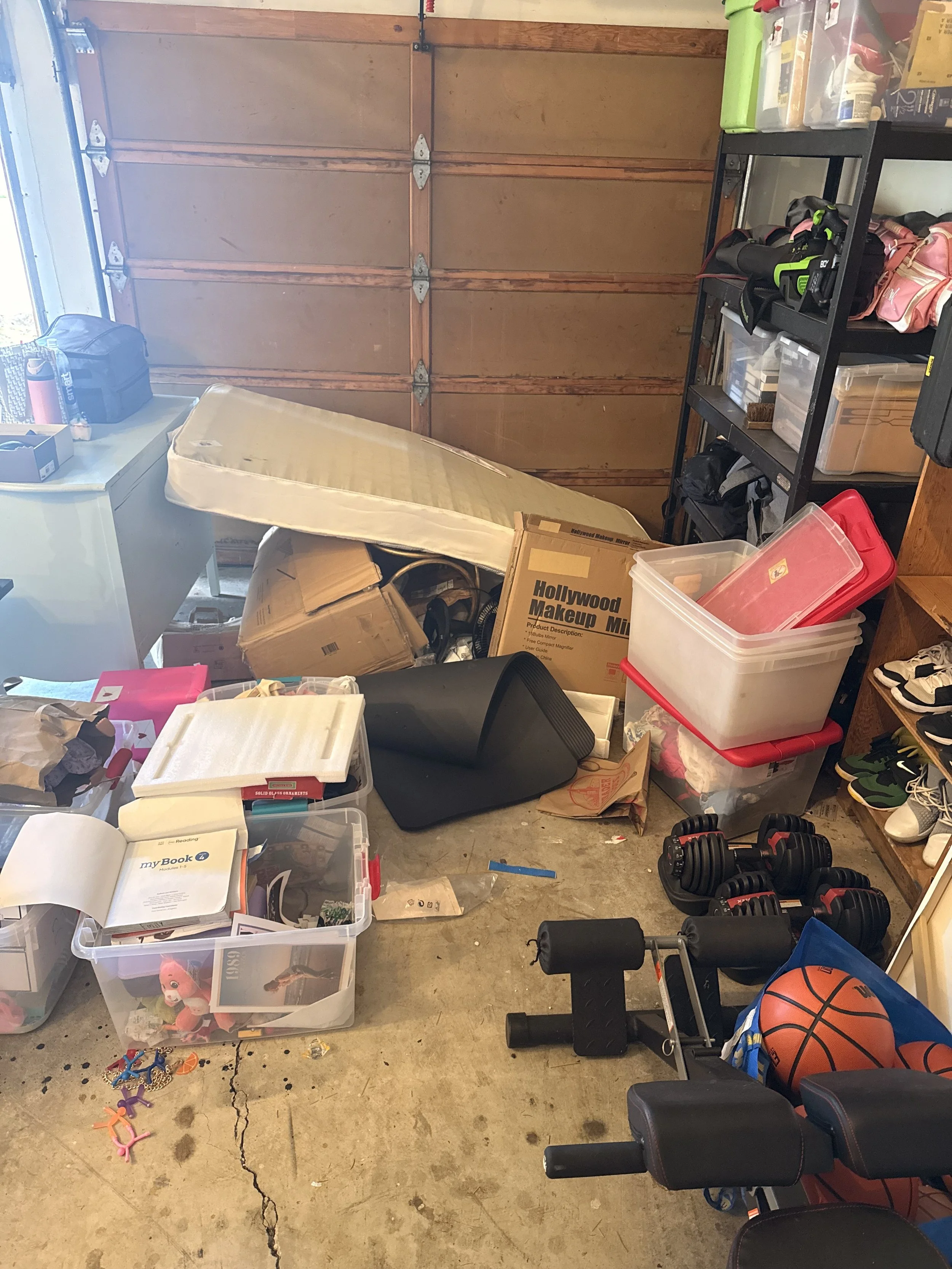 A cluttered garage workspace with shelves filled with tools and boxes, a weight bench, dumbbells, a basketball, children's toys, a cardboard box, and miscellaneous items scattered on the floor and surfaces.