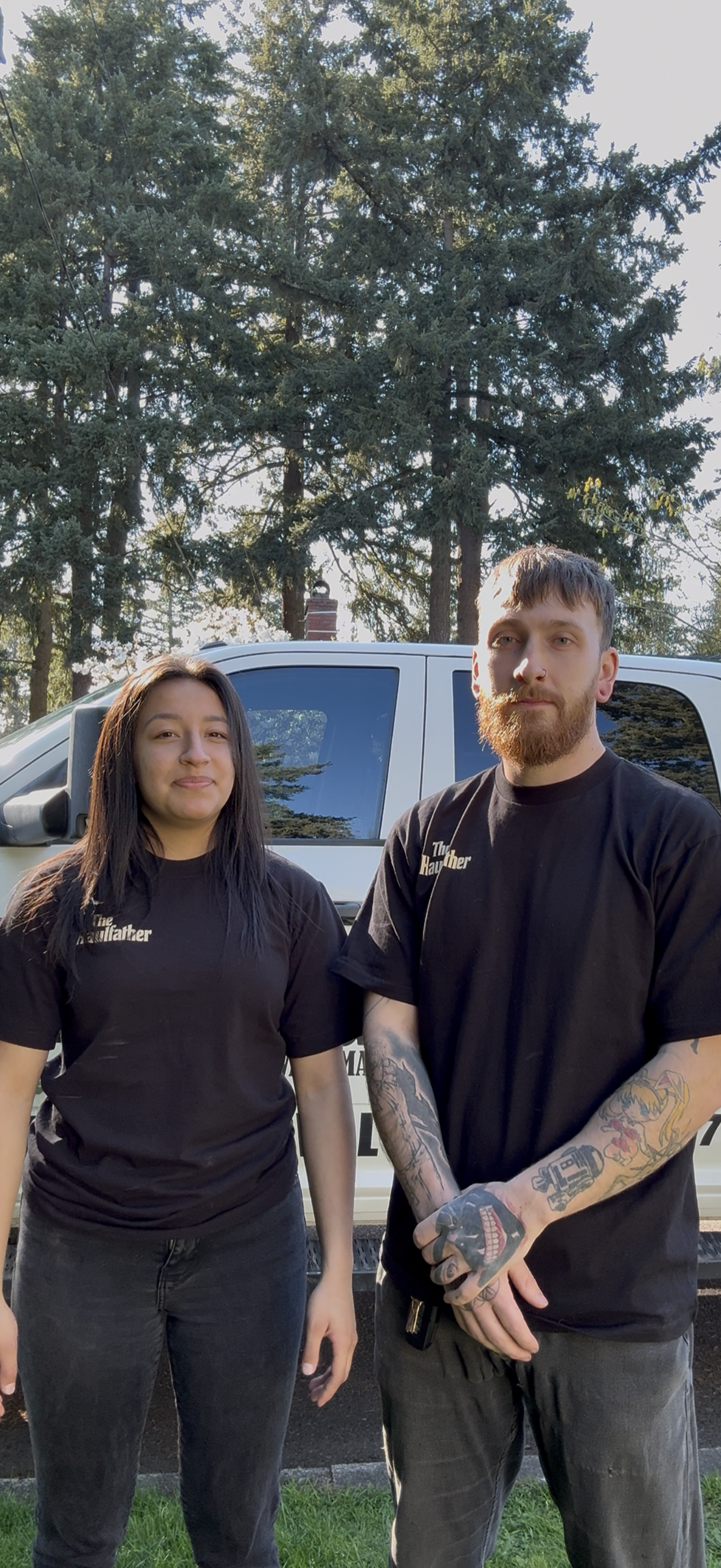 Two young adults, a woman and a man, standing outdoors in front of a white vehicle and tall trees, both wearing black T-shirts with a logo on the front.