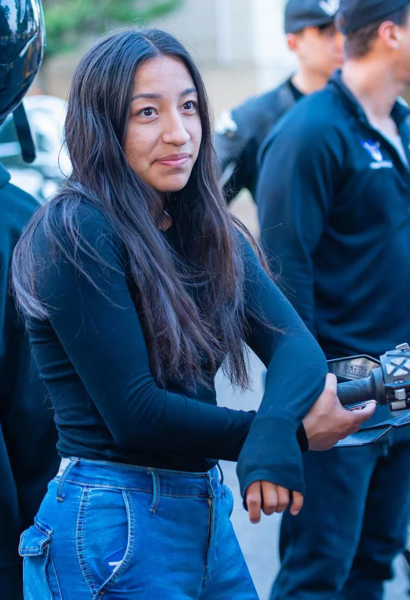 A young woman with long dark hair wearing a black long-sleeve shirt and blue jeans, holding a motorcycle handlebar, standing outdoors with a neutral expression.