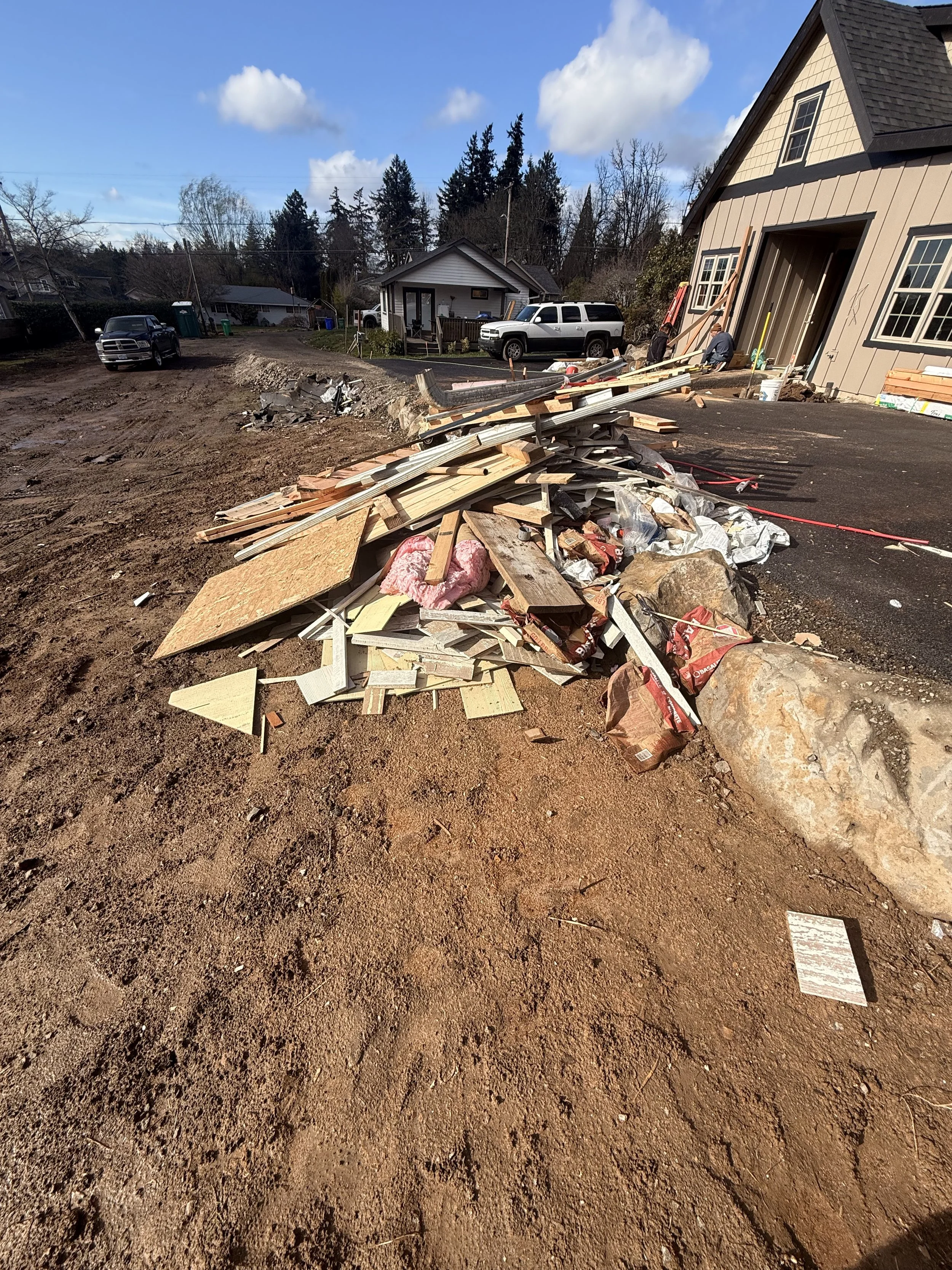 A construction site with dirt and debris, including wood, drywall, rocks, and insulation, in front of a partially built or renovated house with workers. There are parked cars and a small house in the background under a partly cloudy sky.