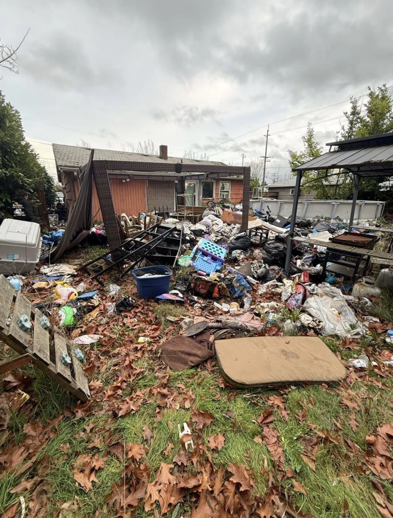 Backyard filled with trash, broken furniture, and scattered debris, leaves covering the ground, cloudy sky overhead.