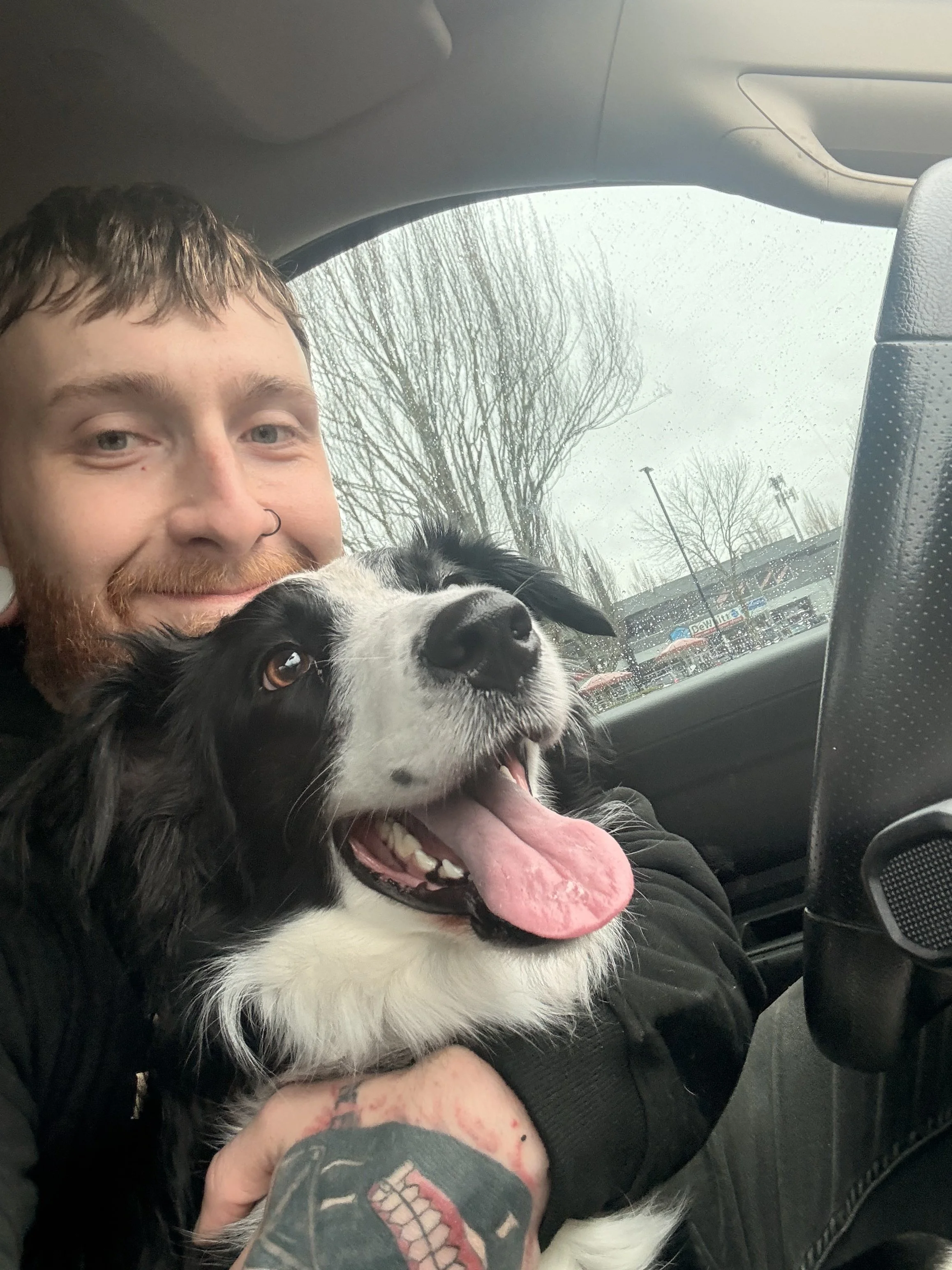 Person with a beard and nose piercing smiling with a black and white dog inside a car. The dog's tongue is out, and wet weather is visible through the window with trees and shop signs in the background.