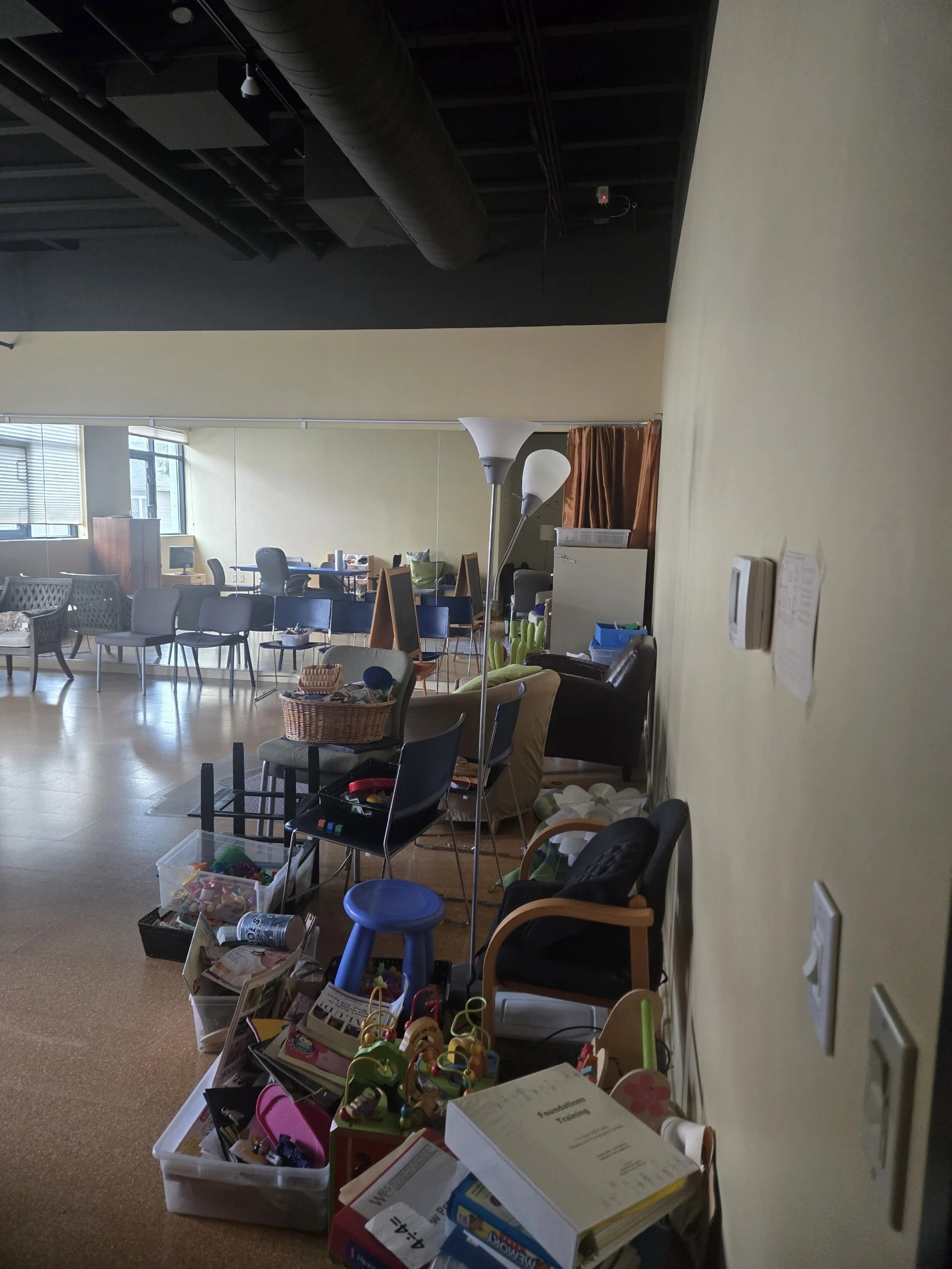 A room with various chairs, tables, and boxes filled with toys, books, and office supplies, with a window and some floor lamps in the background.