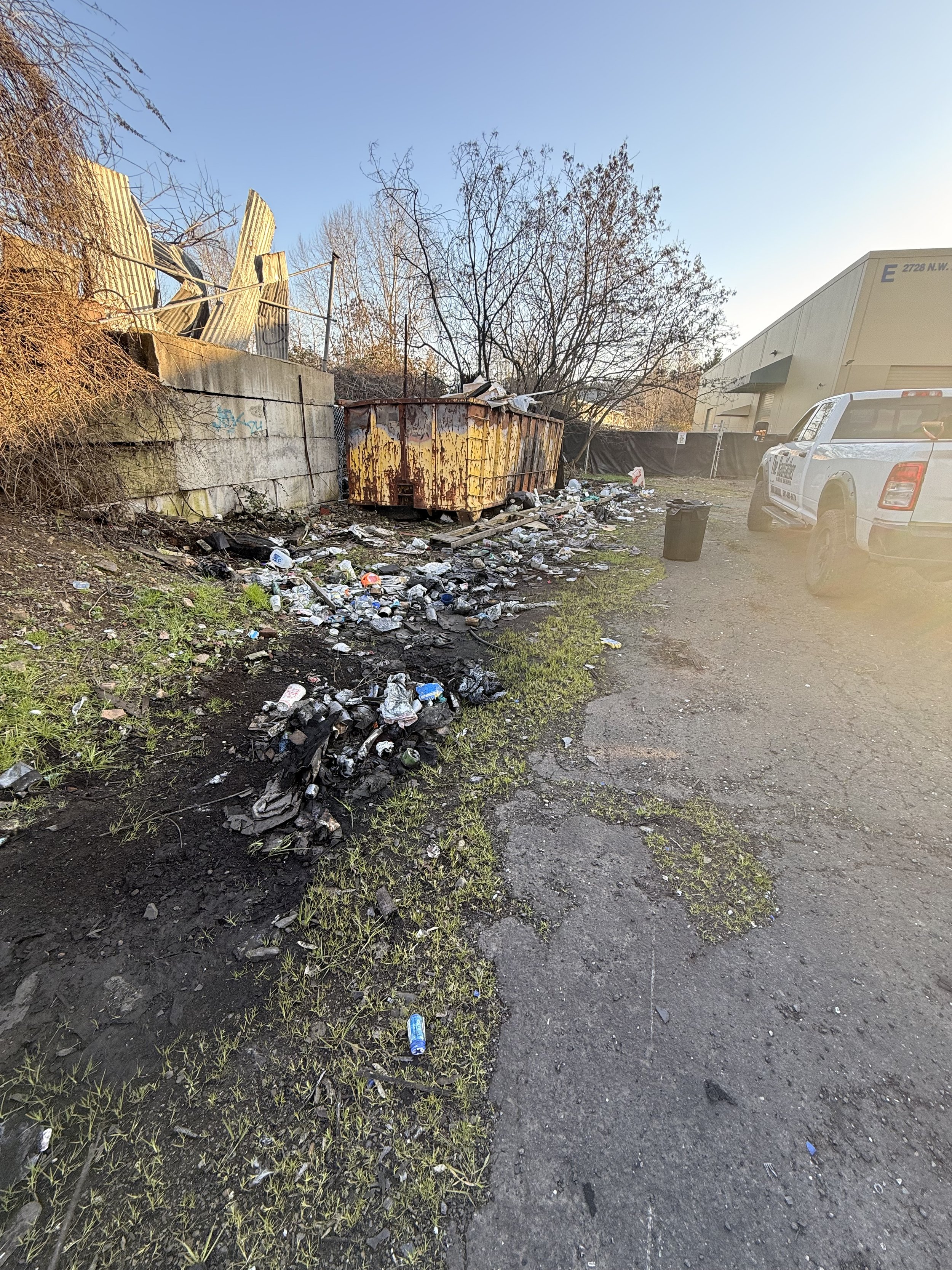 An outdoor scene with a large trash dumpster covered in rust, surrounded by scattered litter, including cans and bottles. There is a concrete wall, leafless trees, a white vehicle, and a warehouse in the background, with the sky clear and blue.