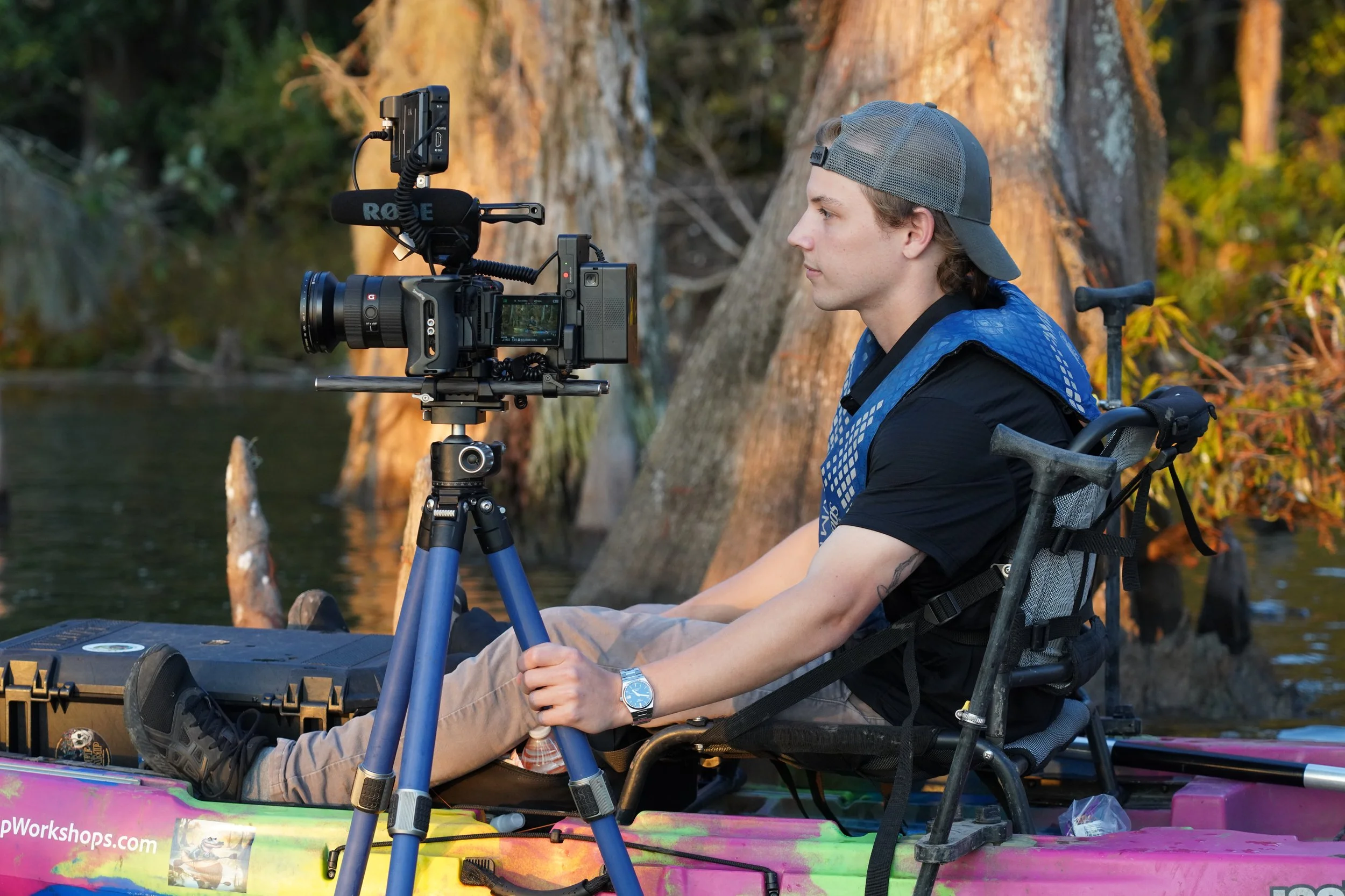 A person sitting in a kayak with a camera mounted on a tripod in front of them, near a body of water with trees in the background.