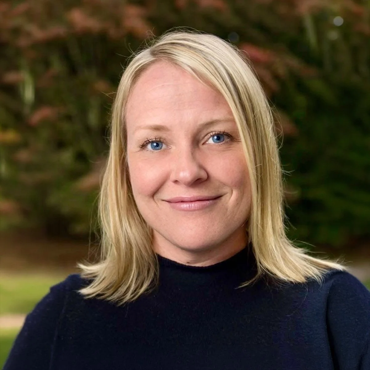 A woman with shoulder-length blonde hair, blue eyes, and a light complexion, smiling outdoors with a blurred background of greenery.