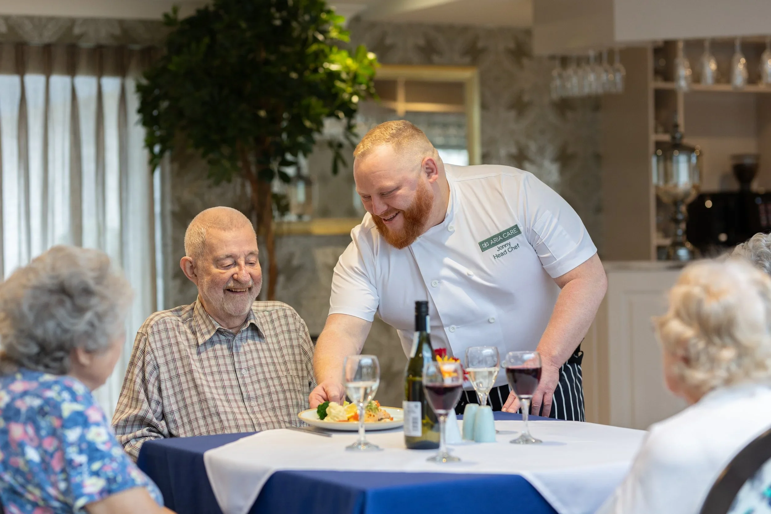 A care home chef serving food to elderly people at a dining table in a care facility.
