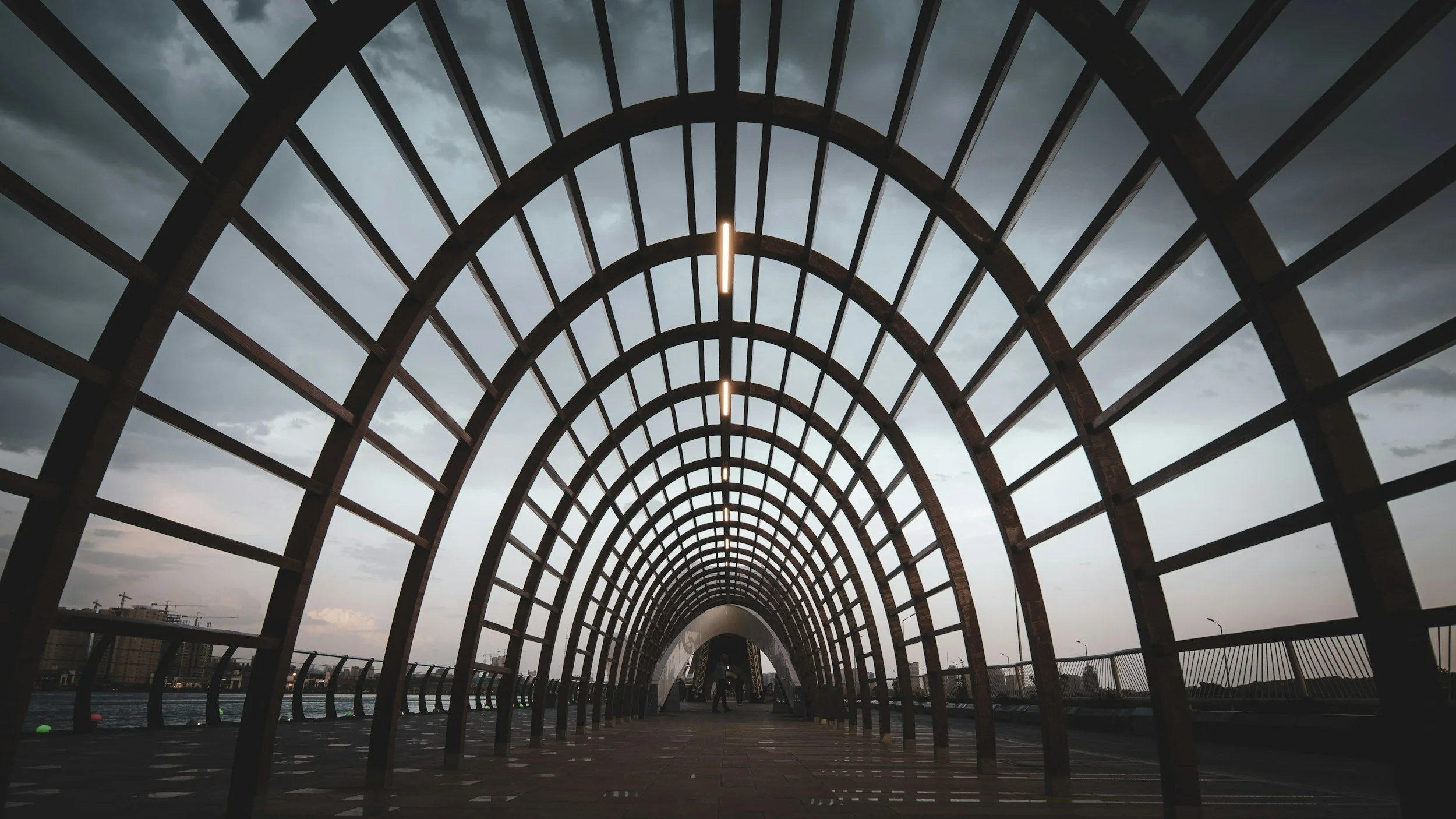 A curved, open-air pedestrian bridge with a metal arch structure and overhead lights, seen from below during cloudy weather.