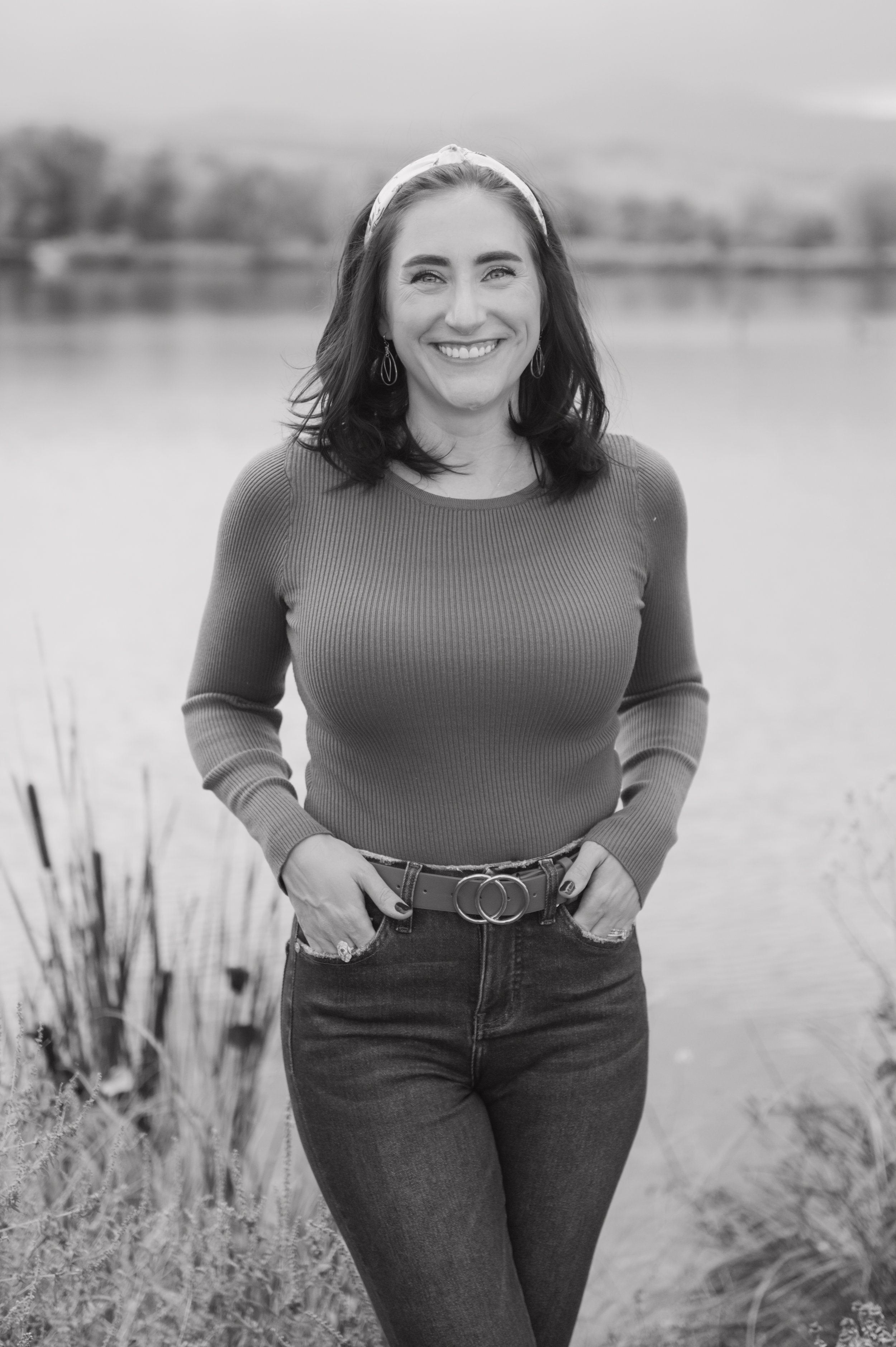 A woman standing outdoors near water with a blurred background of trees, wearing a long-sleeved top, jeans, and a headband, smiling at the camera.