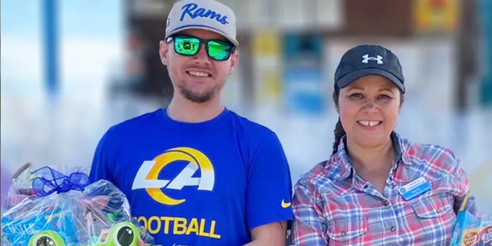 A man and woman smiling inside a store, holding shopping baskets filled with gifts and toys, wearing casual clothes and baseball caps.