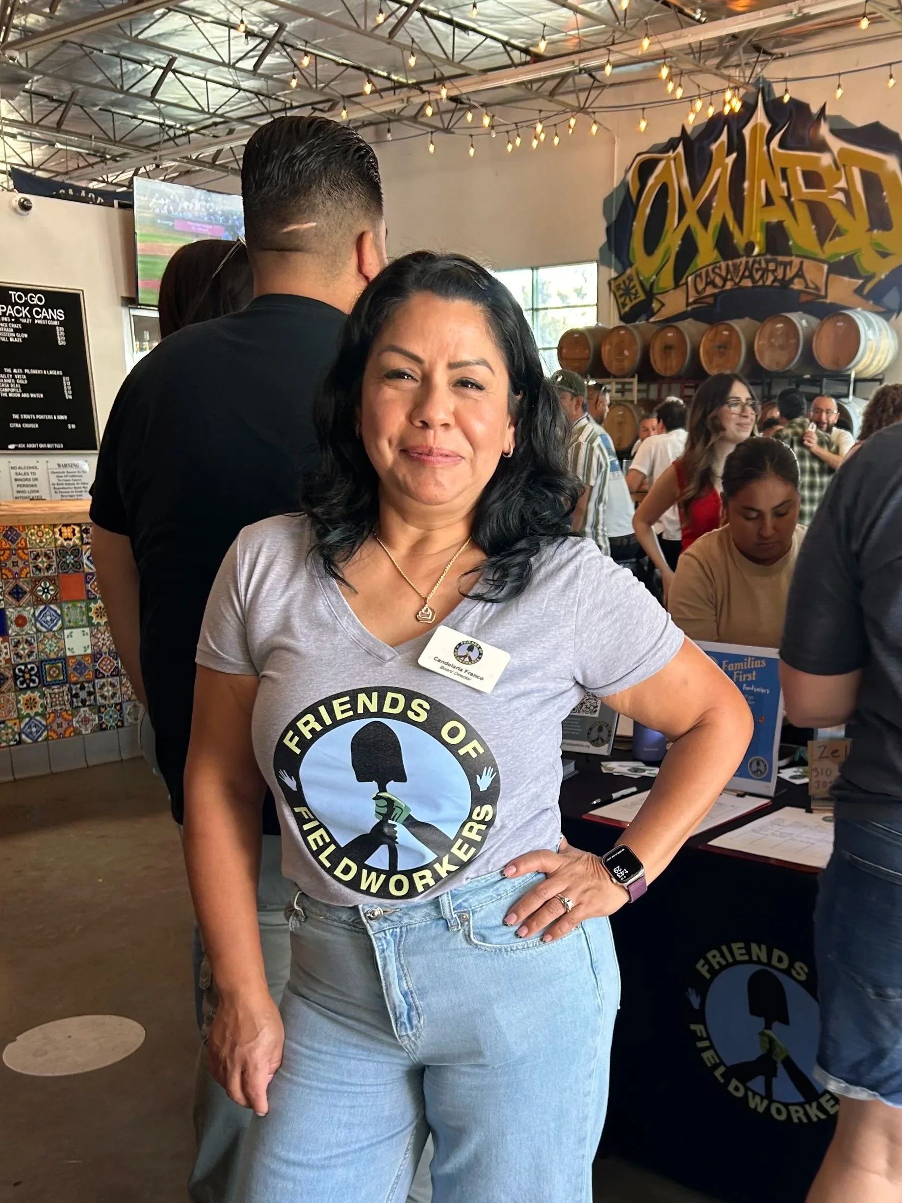 A woman with dark hair, wearing a light gray t-shirt and light blue jeans, standing inside a busy brewery or bar. She has a name tag that reads 'Candelaria Franco' and a logo on her shirt that says 'Friends of Fieldwork Brewers.' She is smiling with 