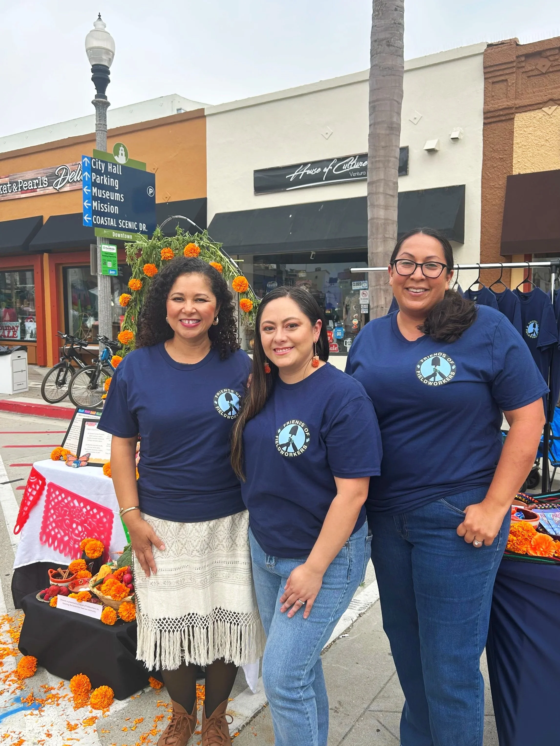 Three women standing outdoors in front of a display table with orange flowers and decorative items, wearing matching blue shirts with a logo that reads 'Friends of Fieldworkers', smiling at the camera. Behind them is a street sign pointing to City Ha