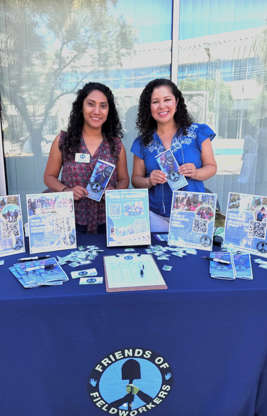 Two women standing behind a table with informational flyers, brochures, and QR codes, representing 'Friends of Fieldworkers,' in an outdoor setting with a glass window and trees reflected behind them.