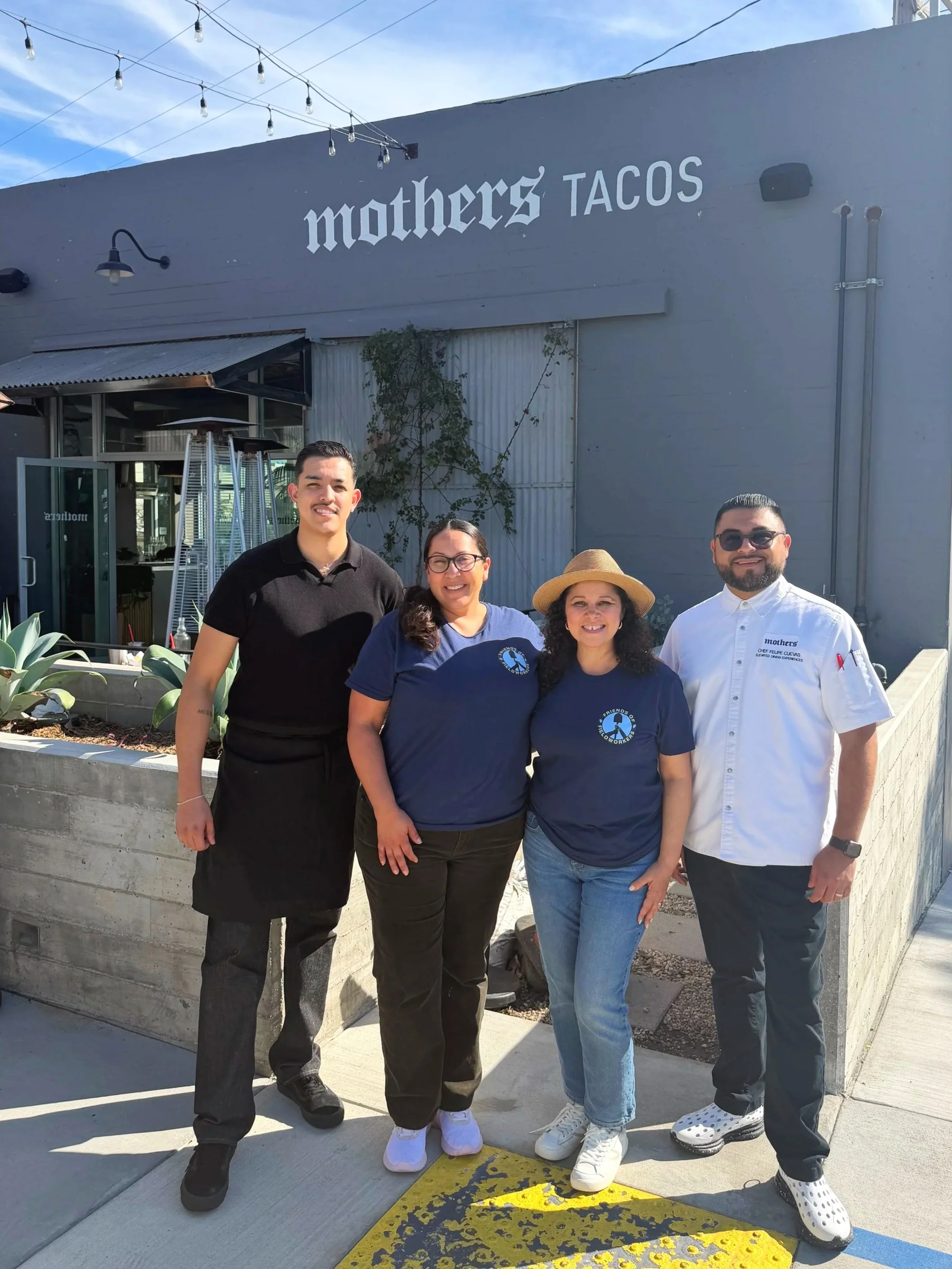 Four people standing in front of a restaurant called 'mothers TACOS'. They are smiling and pose for the photo, with outdoor string lights above and a small garden bed with succulents in front.