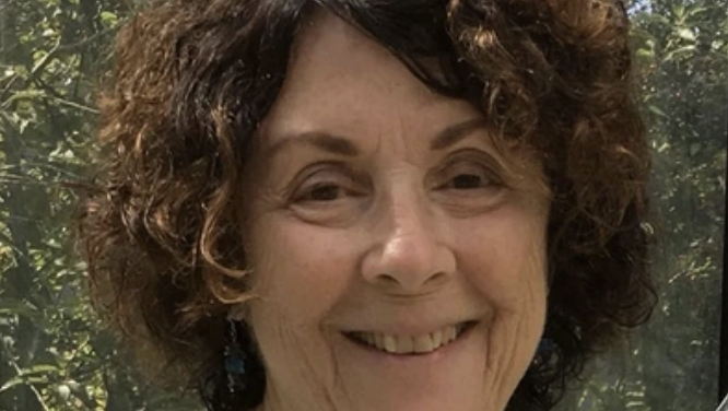 Close-up of a smiling woman with curly brown hair outdoors, with green foliage in the background.