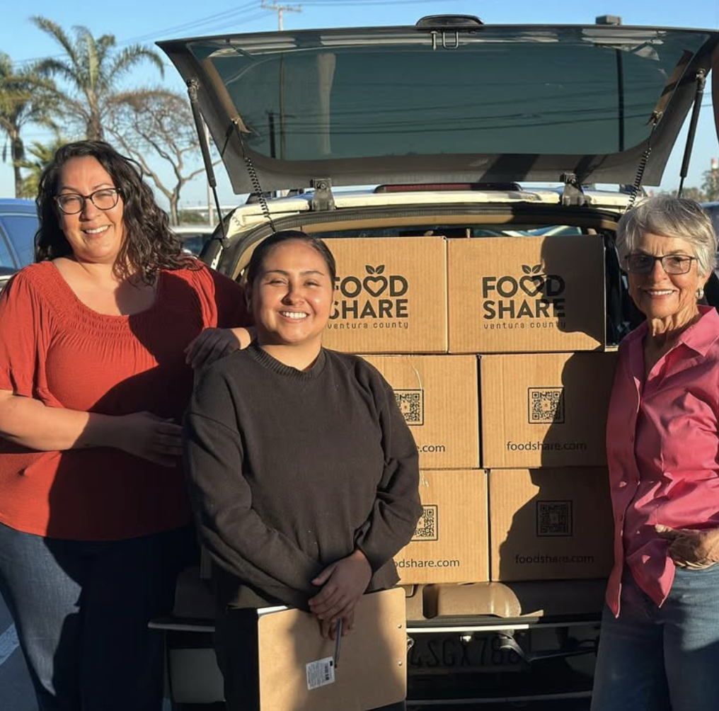 Three women standing in front of an open trunk filled with boxes labeled Food Share, Ventura County, smiling at the camera.