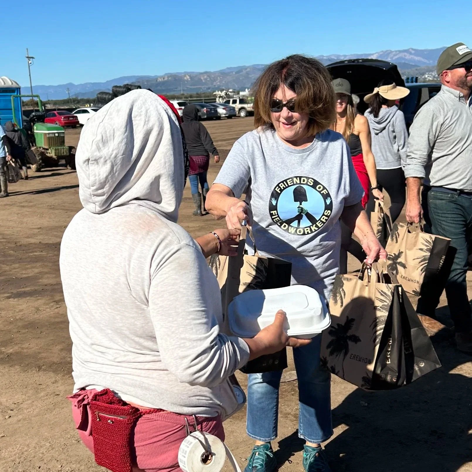 A group of people outdoors at a festival or event, with women holding tote bags and cans, some wearing hats and sunglasses, in a sunny open area with mountains in the background.
