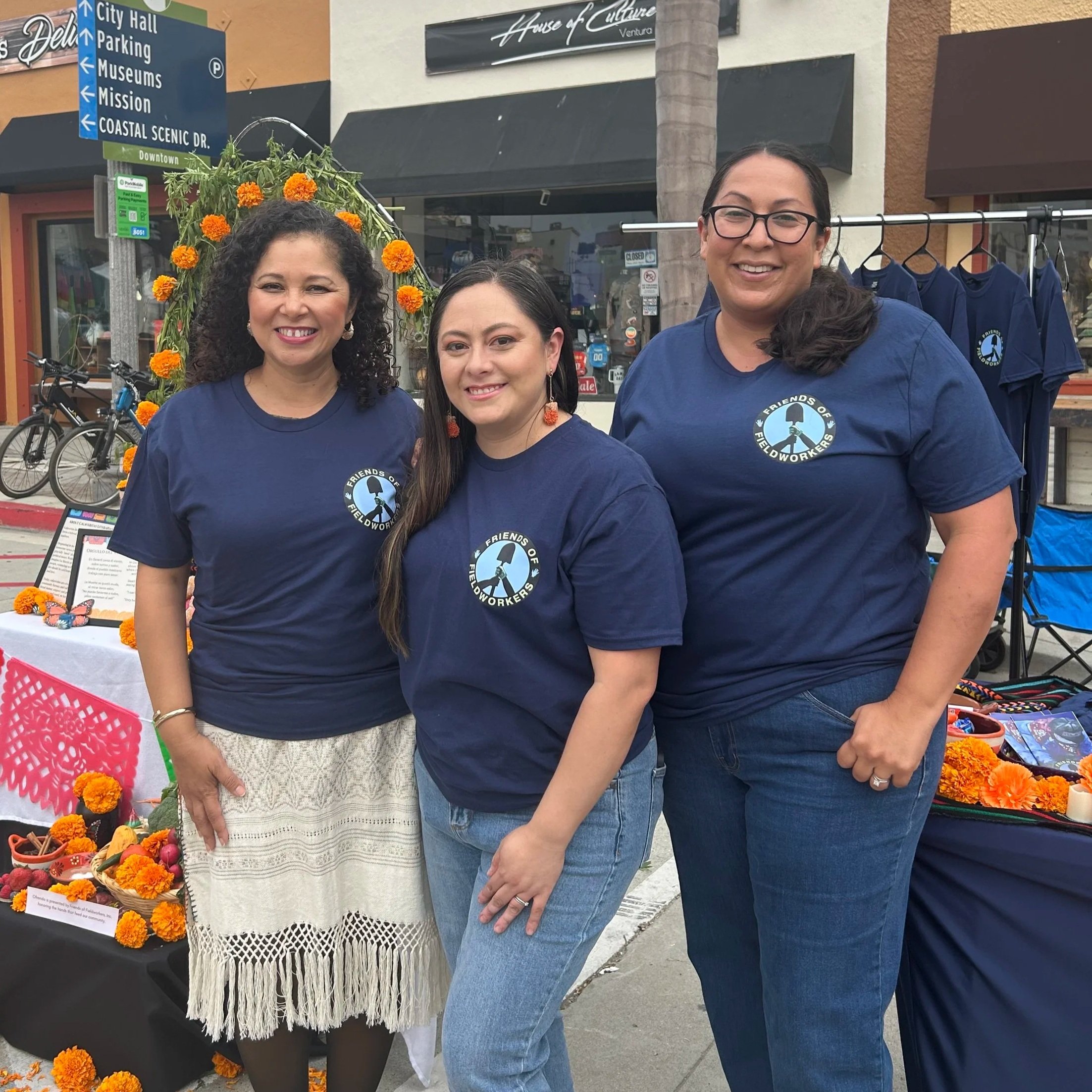 Three women wearing matching blue t-shirts with a logo, standing together outdoors in front of a display of marigold flowers and a sign labeled "Friends of Fieldworkers." The background includes a storefront, bicycles, and a street sign pointing to City Hall, Parking, Museums, Mission, and Coastal Scenic Drive.