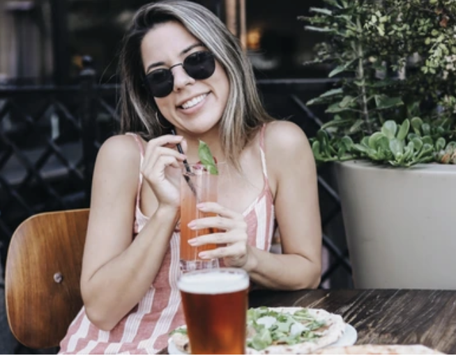 Young woman in sunglasses smiling while drinking a cocktail at an outdoor restaurant, with a pizza and a glass of beer on the table, and potted plants in the background.