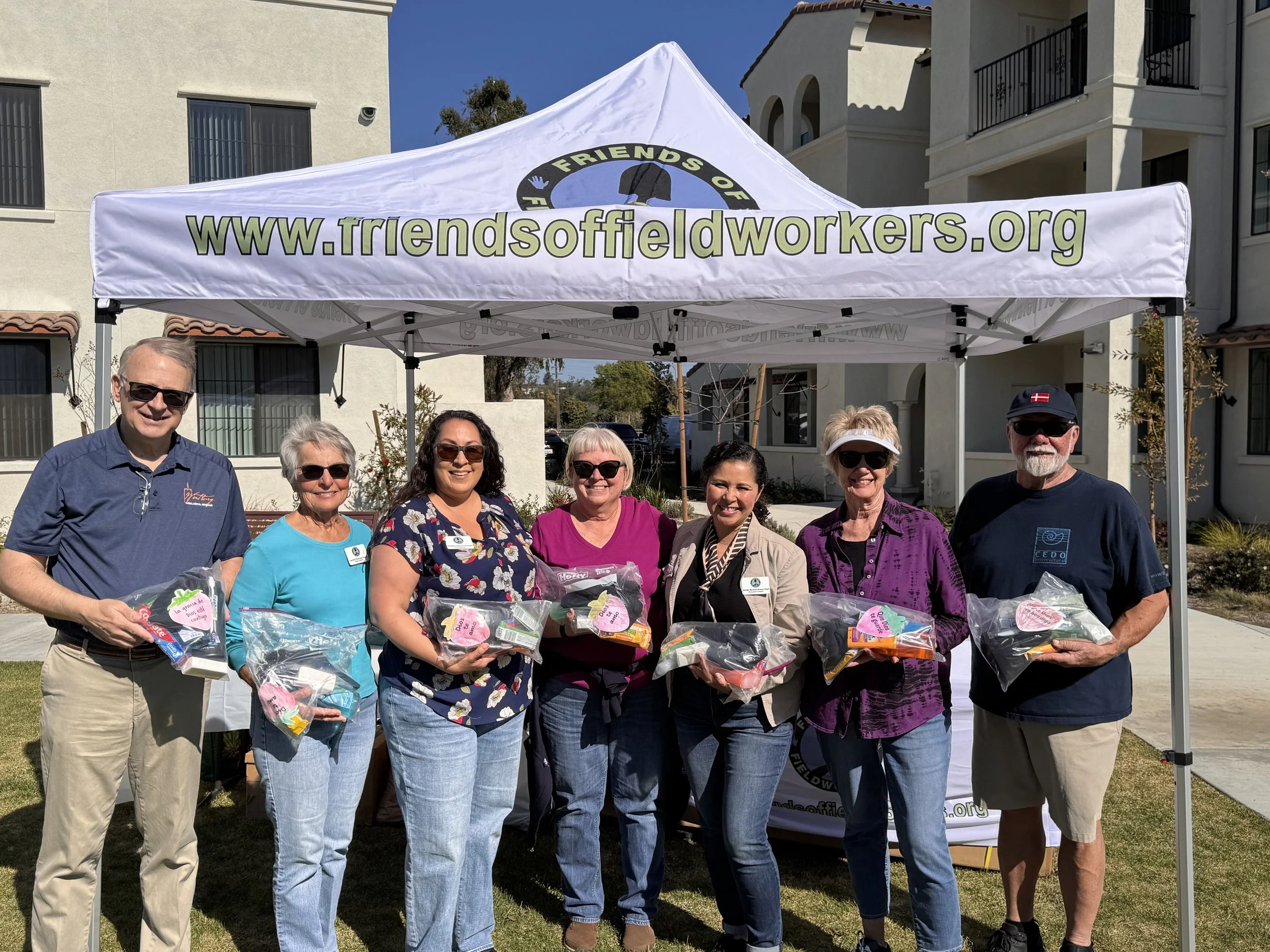 A group of people standing under a white canopy with the website 'www.friendsoffieldworkers.org' printed on it, holding clear plastic bags filled with colorful items, in an outdoor residential area.