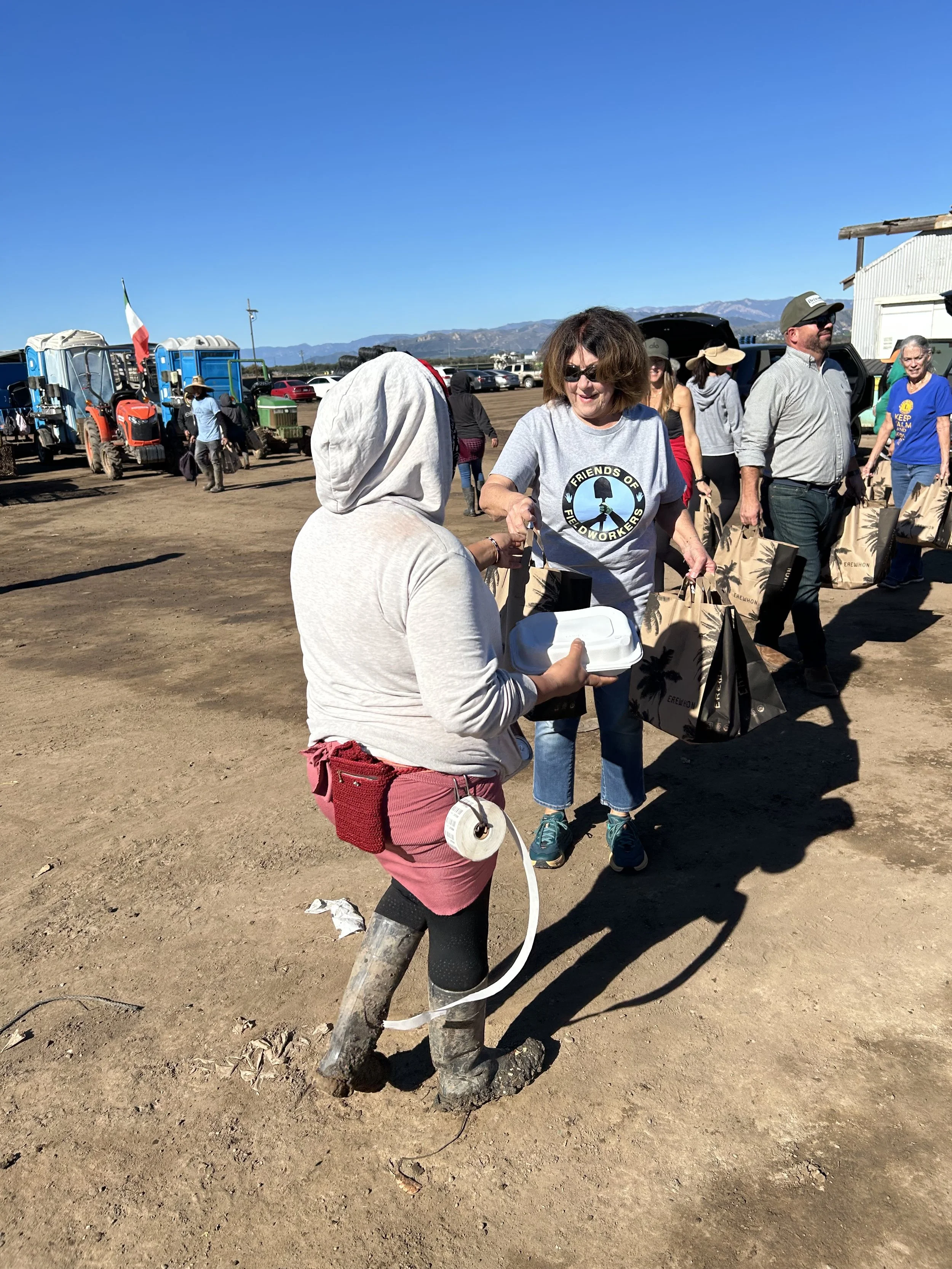 People gathered outdoors near tractors and vehicles under a clear blue sky, engaging in conversation, with a mountain range in the background.