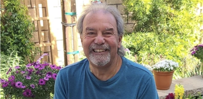 Smiling older man with gray hair and beard sitting in a garden with flowers and greenery.