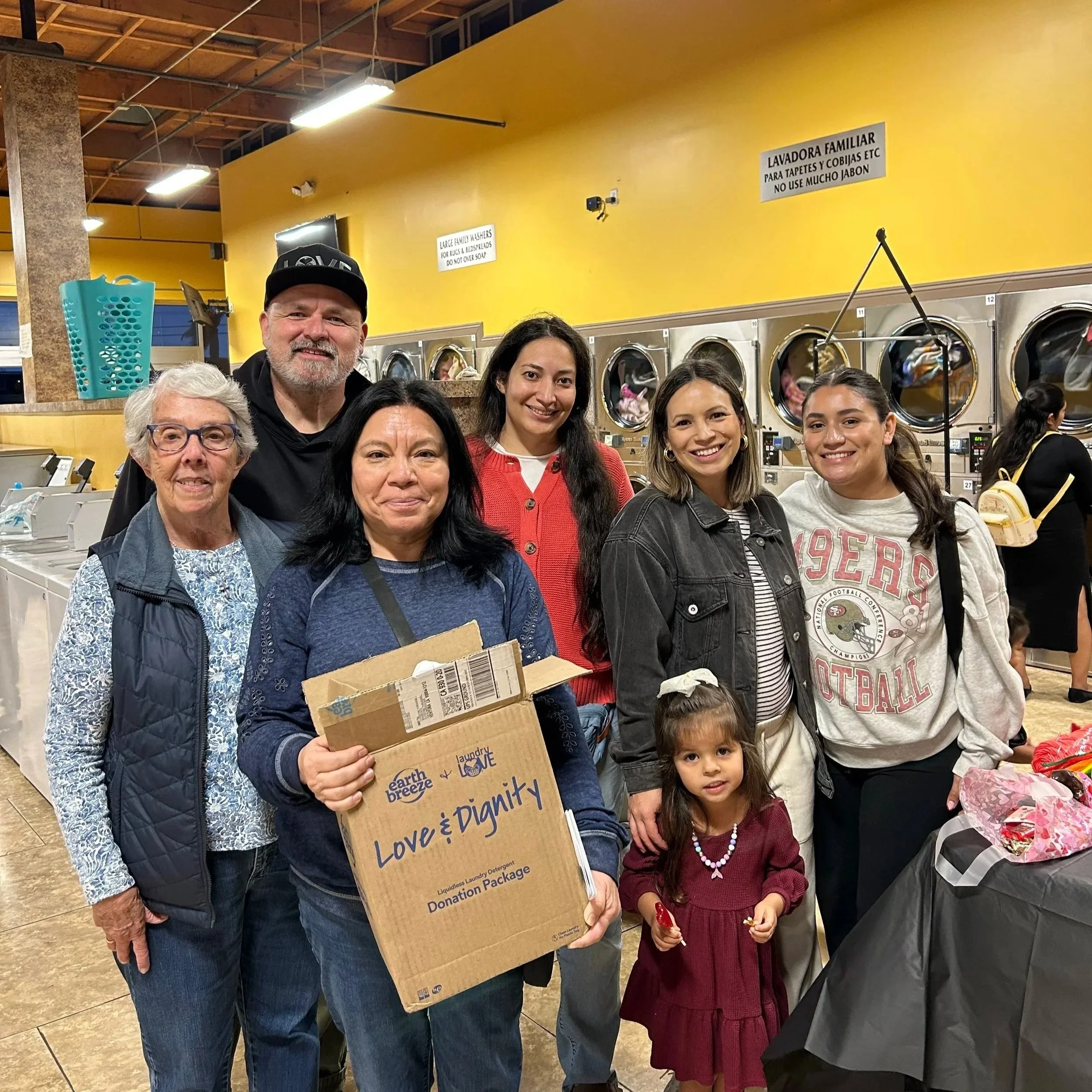 Group of diverse people smiling at a laundromat, some holding a cardboard box that reads 'Love & Dignity' and 'Donation Package', with washers and yellow walls in the background.