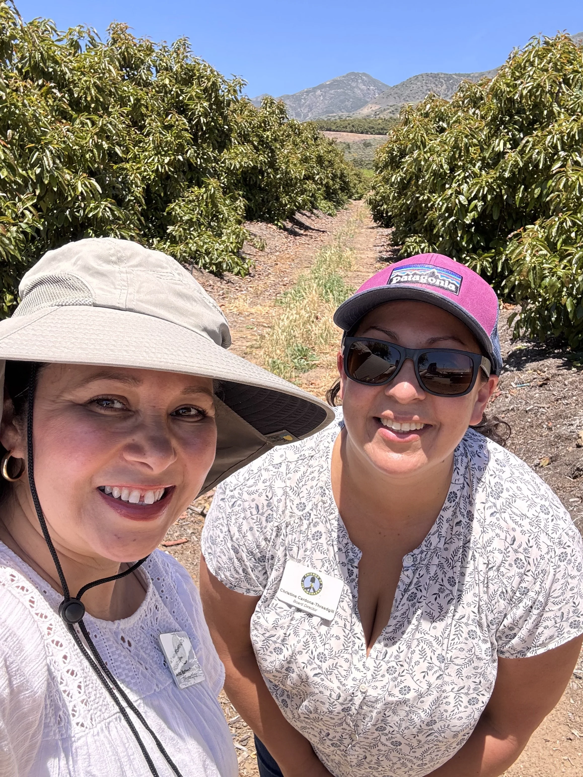Two women smiling for a selfie outdoors, with rows of crops and mountains in the background, sunny weather.