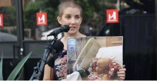 A woman speaking into a microphone outdoors, holding a photo of a young child in a hospital bed with medical equipment.