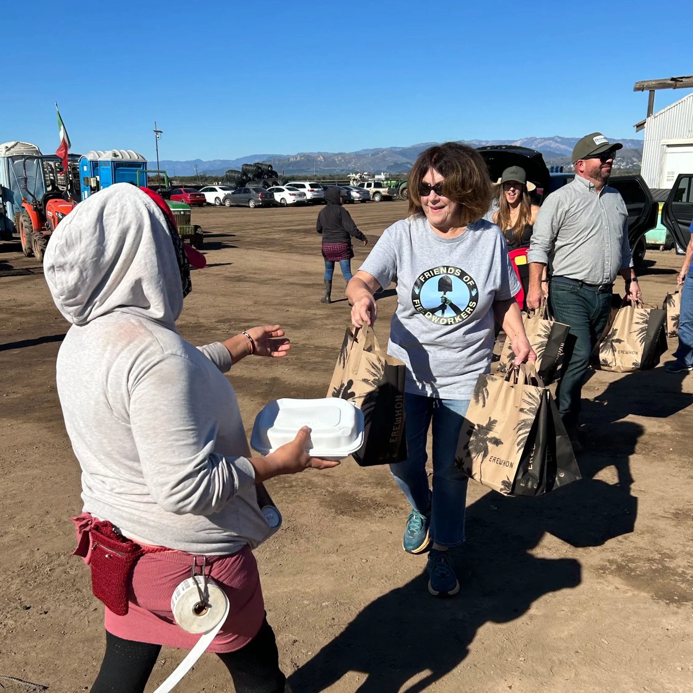 People outdoors at an event, with cars and mountains in the background, some are smiling and carrying bags, while a person in the foreground is handing a box to someone else.