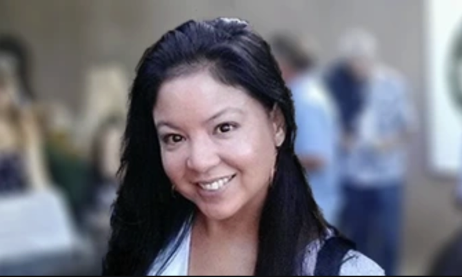 A woman smiling indoors with blurred background of people and furniture.