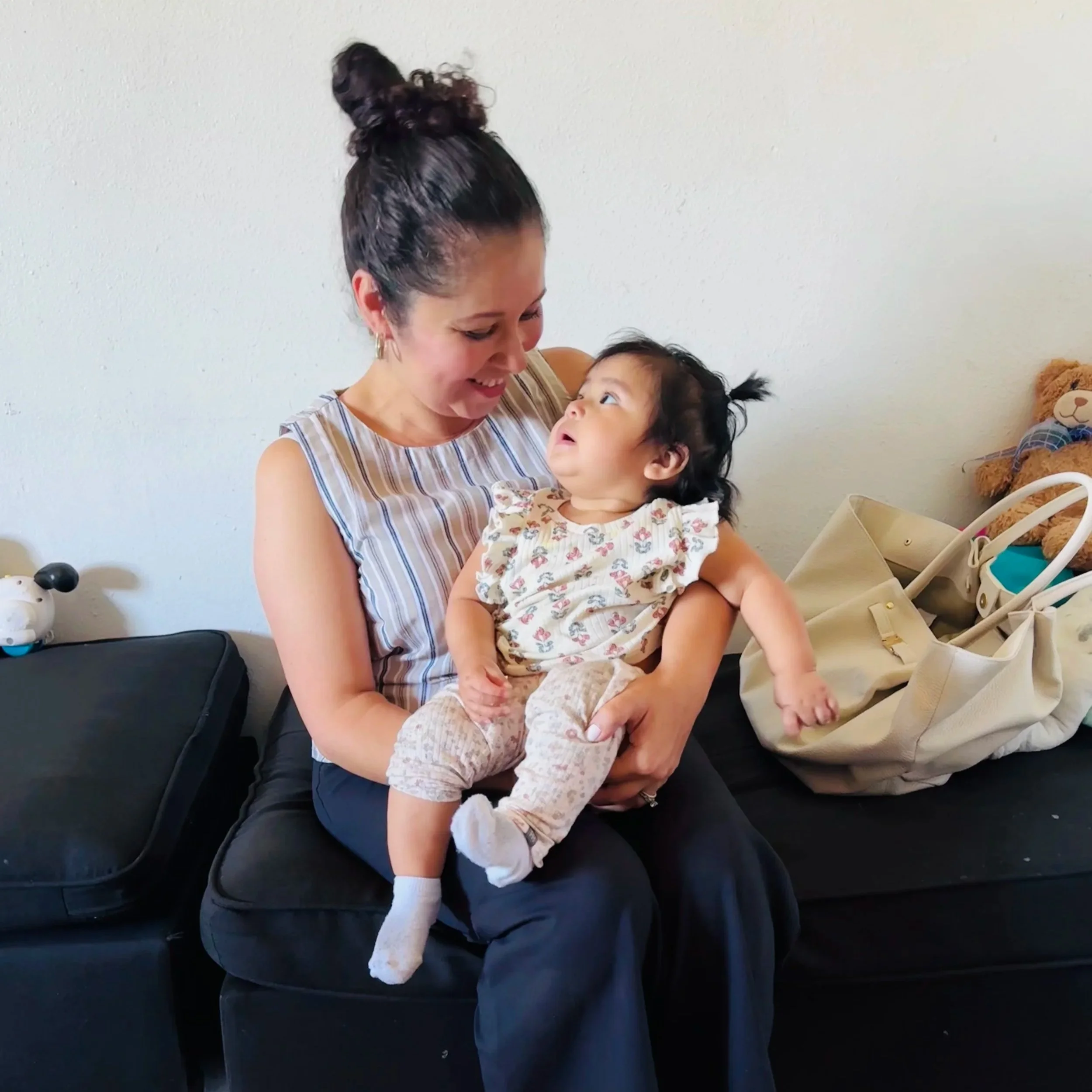 A woman and a young girl sit together on a dark couch, smiling and looking at each other affectionately. The woman has dark hair styled in a bun and is wearing hoop earrings and a striped dress. The girl has dark curly hair and is wearing a floral dress with white socks.