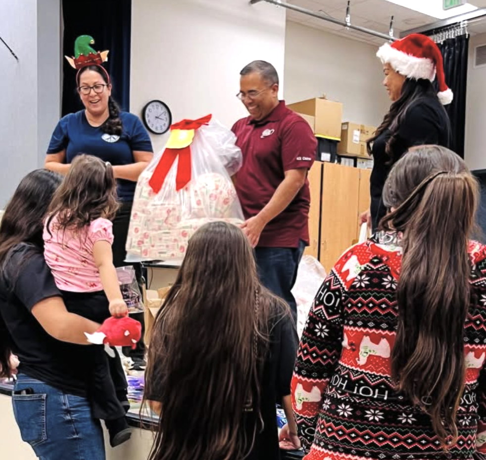 People gathered around a table exchanging a large gift basket, celebrating during a Christmas-themed event, with some wearing holiday hats and sweaters.