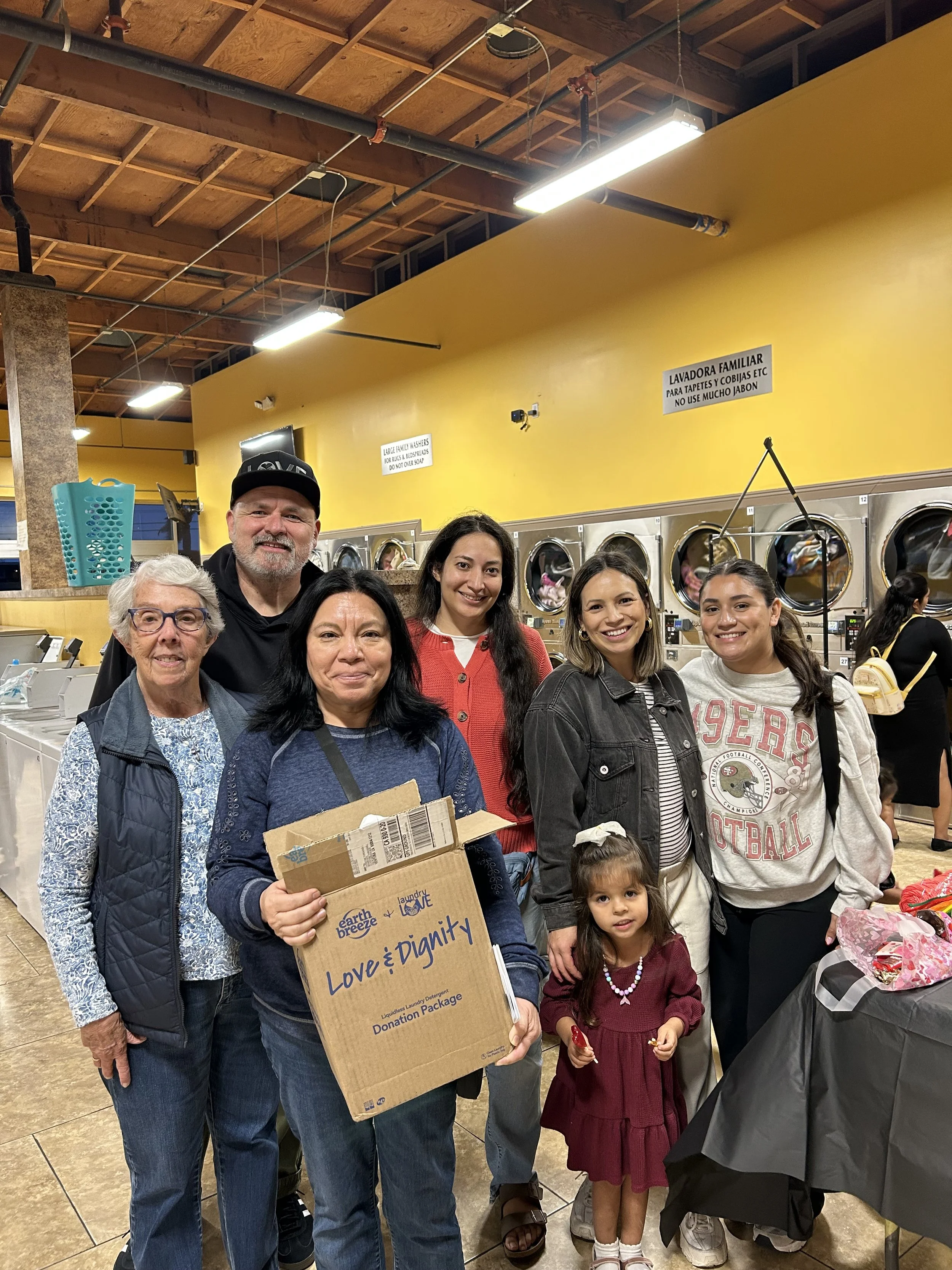 Group of diverse people, including children and adults, standing in a laundromat with yellow walls and washing machines in the background, some holding boxes for donation, smiling at the camera.
