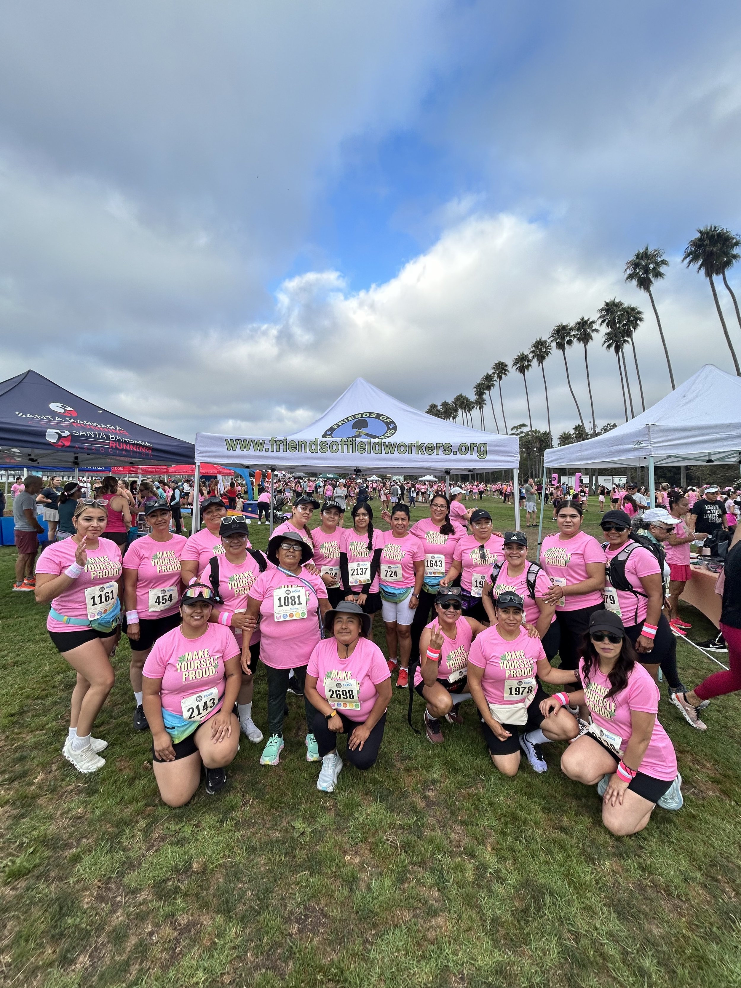 A group of people wearing pink shirts with the message 'Make Yourself Proud' at an outdoor charity run event. Some are kneeling in front, and others are standing behind them, with tents and more people in the background. The sky is partly cloudy with