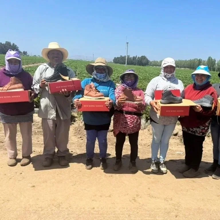 Group of seven women standing in a field, each holding a box with shoes inside, wearing hats and masks, with a clear blue sky and green crops in the background.