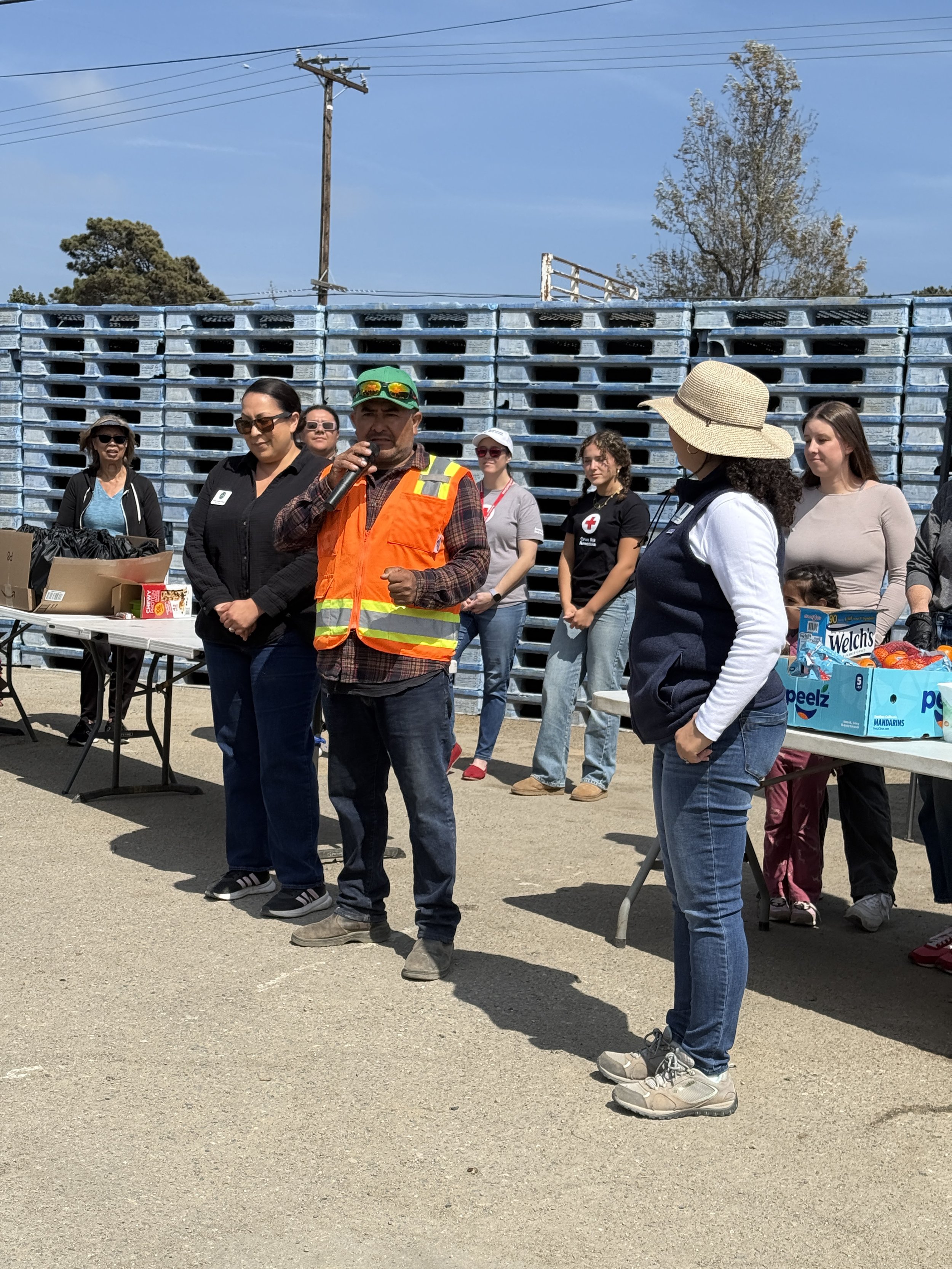 A group of people standing outdoors at a community event, with tables of supplies, behind a tall stack of blue pallets and a wooden utility pole. One man in an orange safety vest is speaking into a microphone.