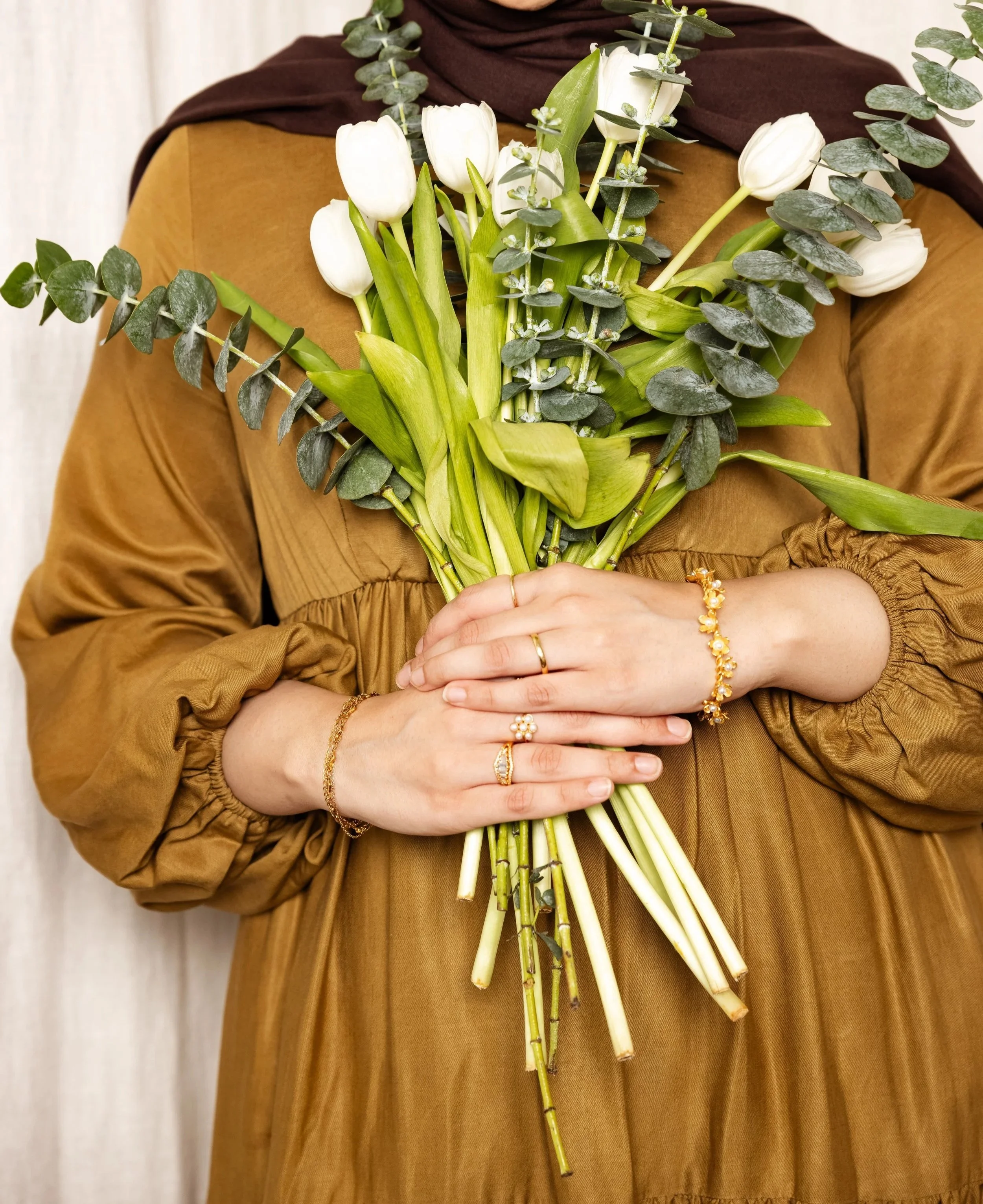 A person wearing a brown dress and gold jewelry holding a bouquet of white tulips and greenery.